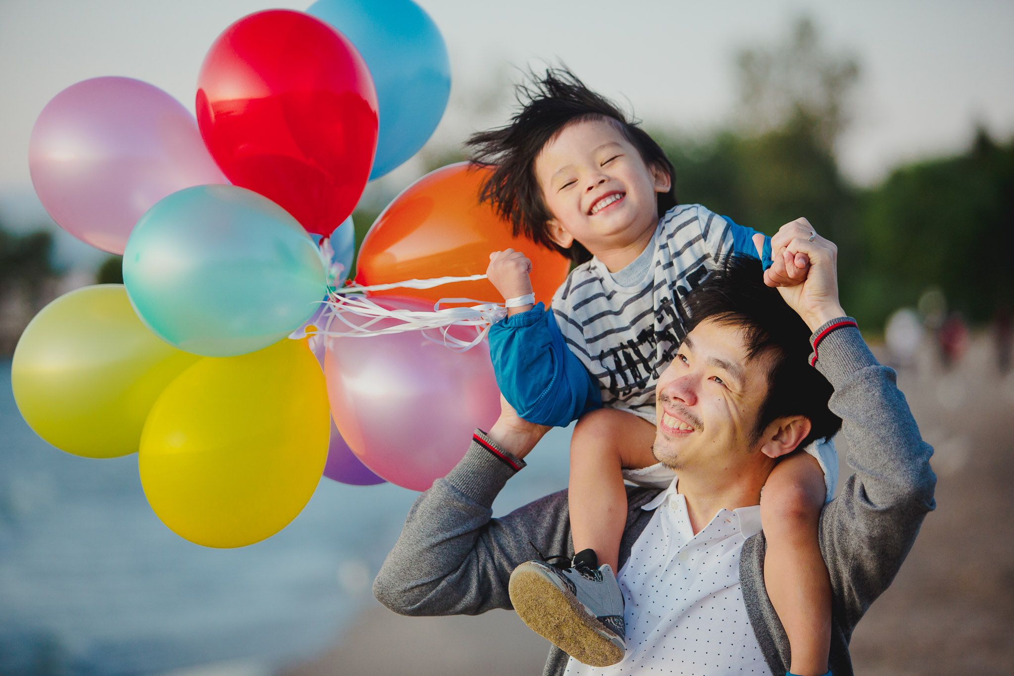 Two people share a joyful moment outdoors with colorful balloons near the water on a sunny day.