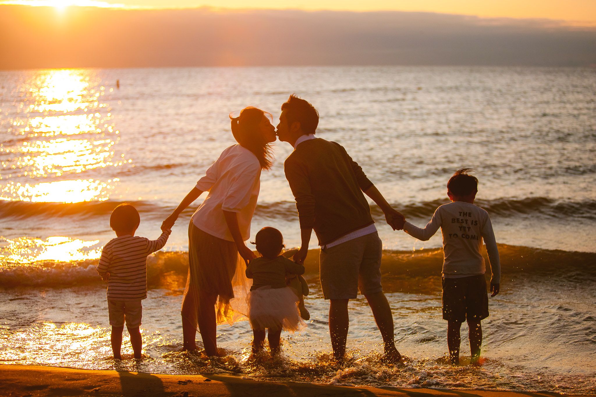 Silhouetted family stands together on beach at sunset with ocean waves and golden light creating magical moment.