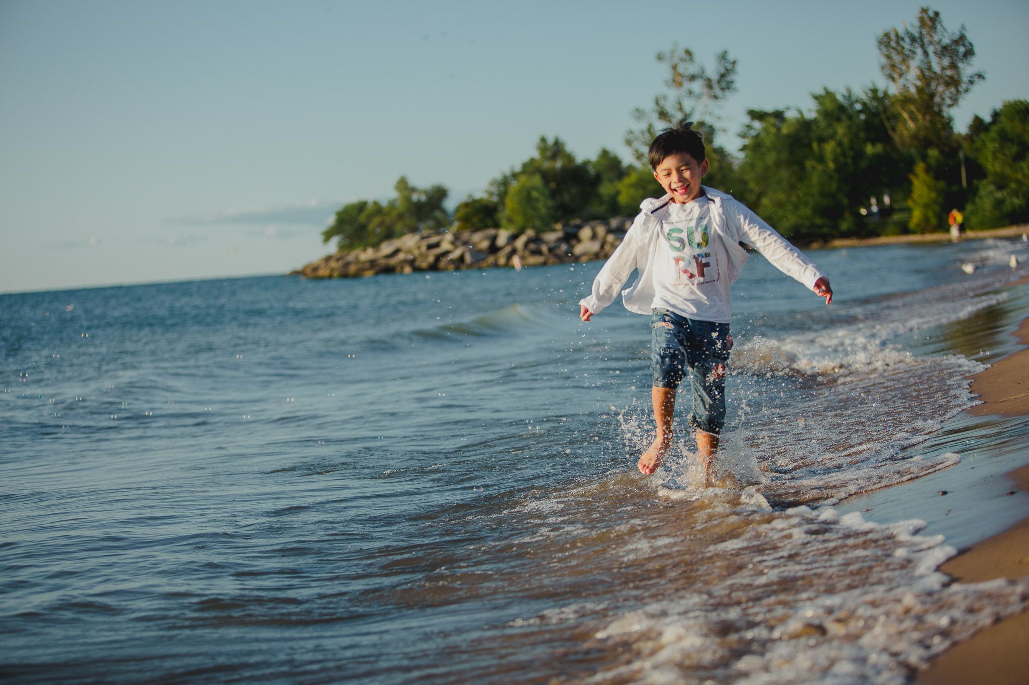Child running and splashing in shallow ocean water at beach during summer day with trees in background.