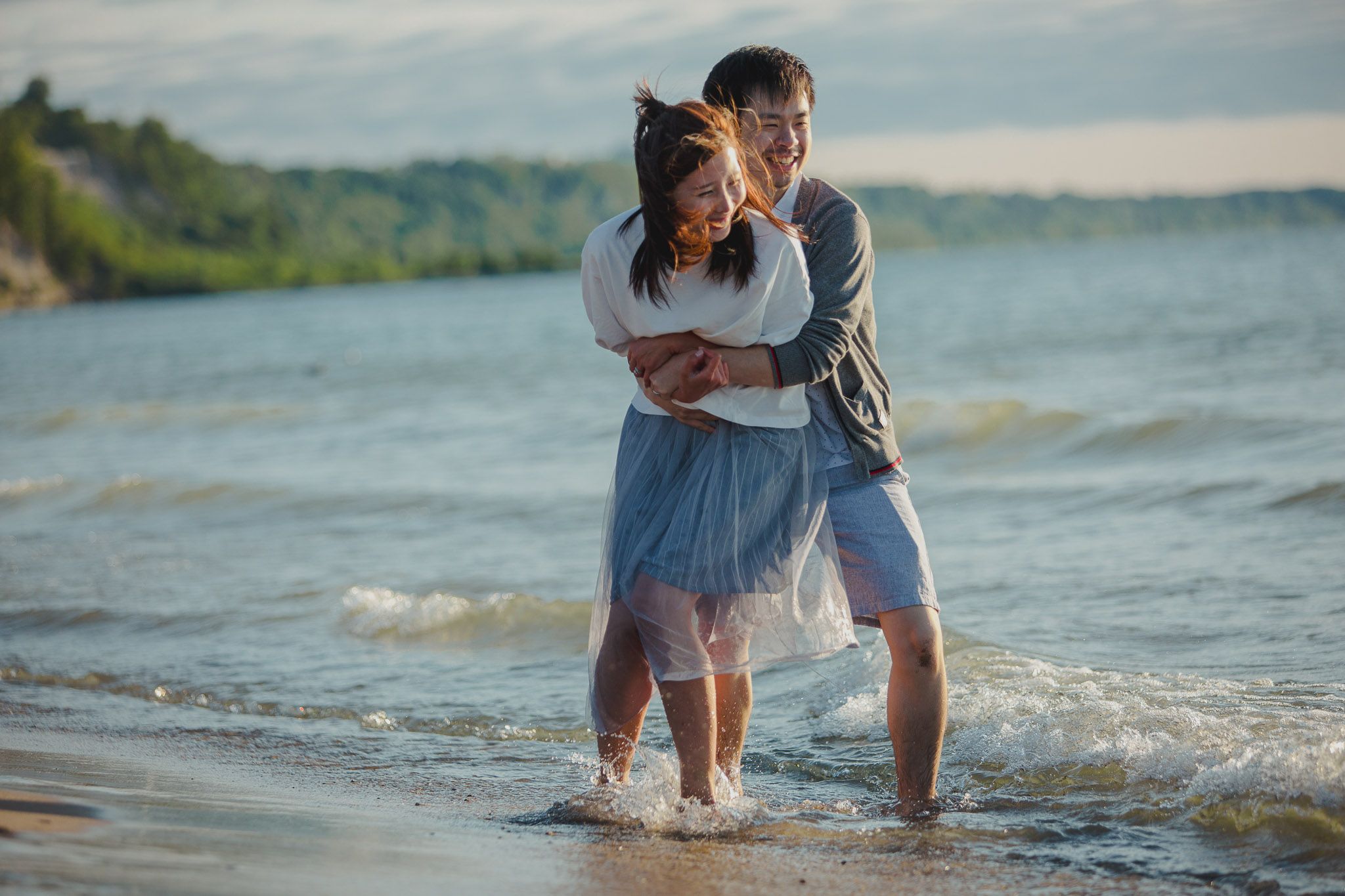 Couple embracing while standing in shallow water at lakeside with green hills in background.