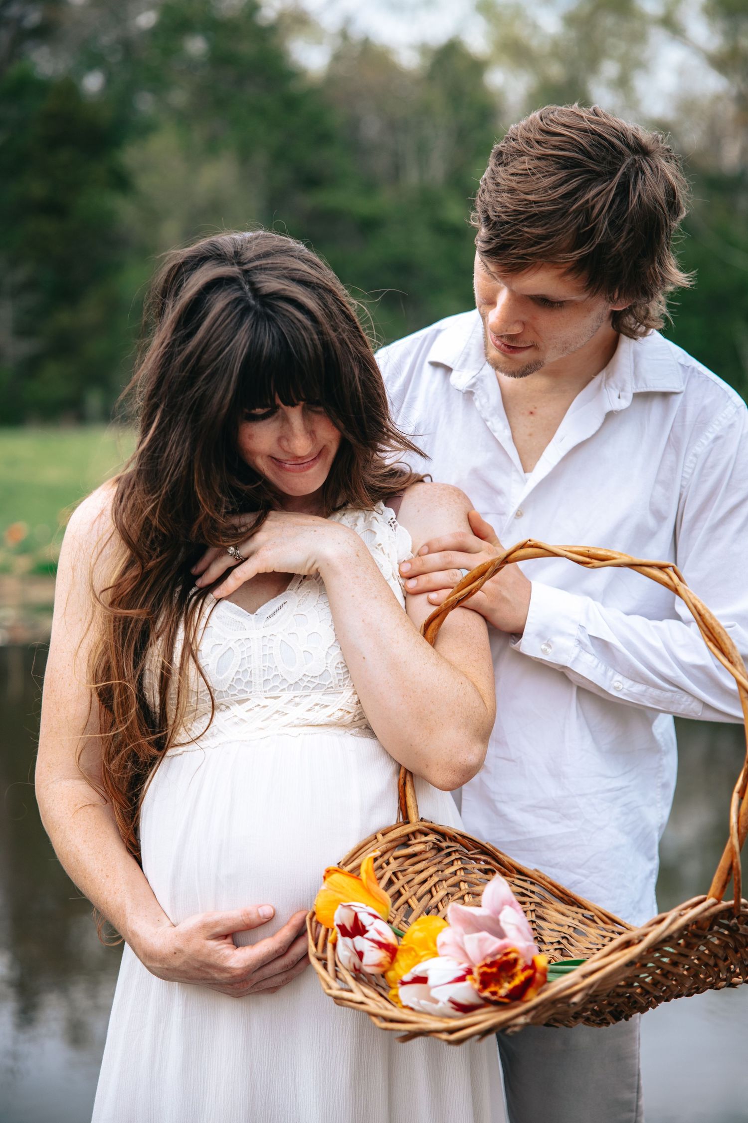 A sweet outdoor maternity photoshoot scene by a pond with a white dress and picnic basket.