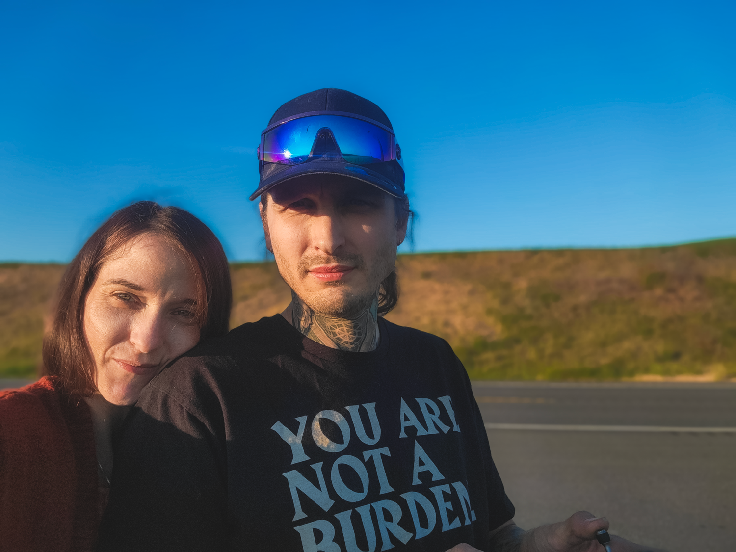 Couple taking outdoor selfie against blue sky and rolling hills backdrop.