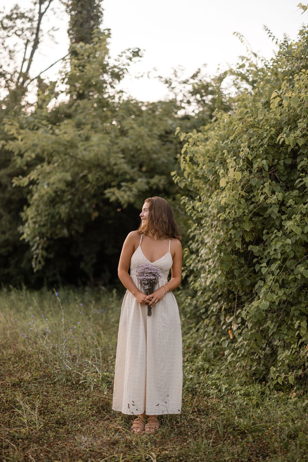 A person in a white summer dress stands in a grassy outdoor setting with greenery in the background.