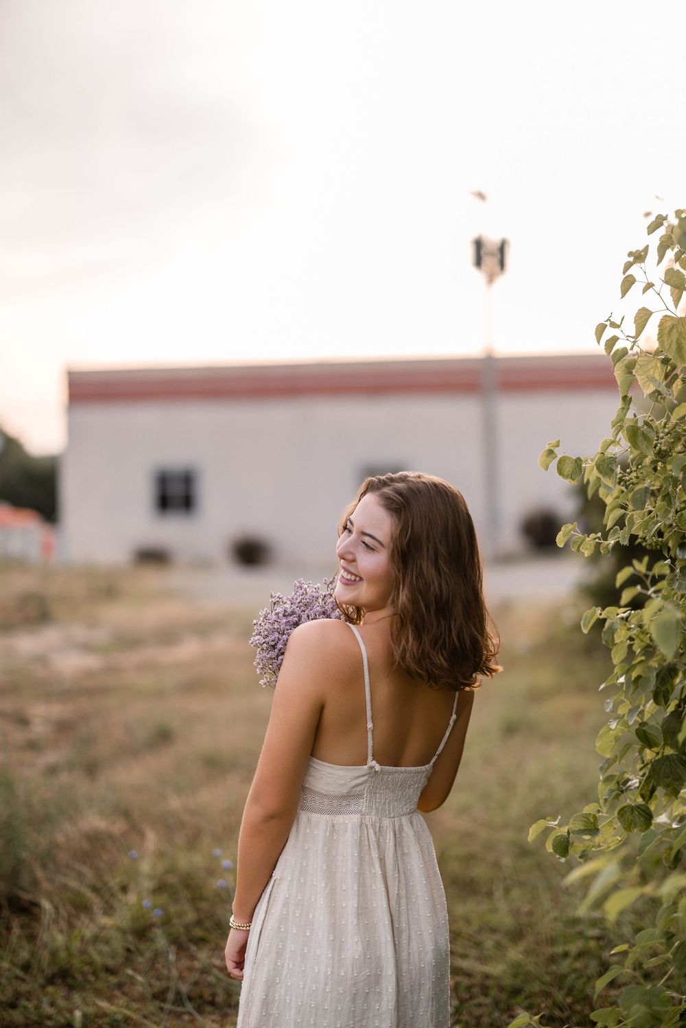 A person in a white summer dress stands in a field at sunset with a butterfly floating nearby while looking over their shoulder.