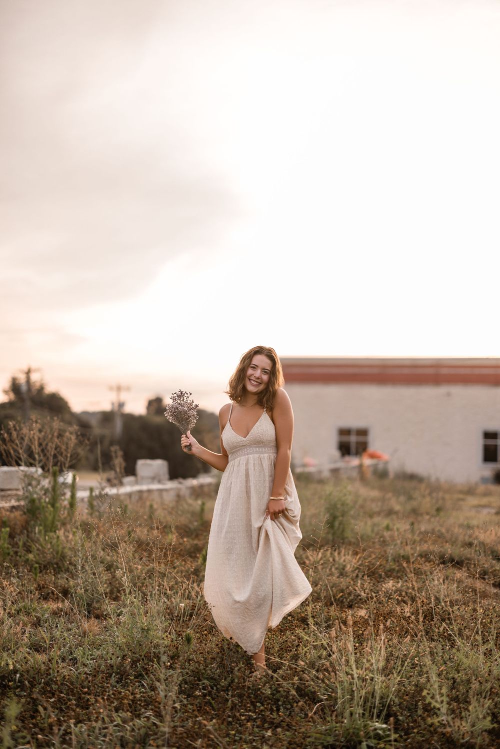 A person wearing a flowing white dress walks through a rustic field at sunset creating a dreamy summer scene.