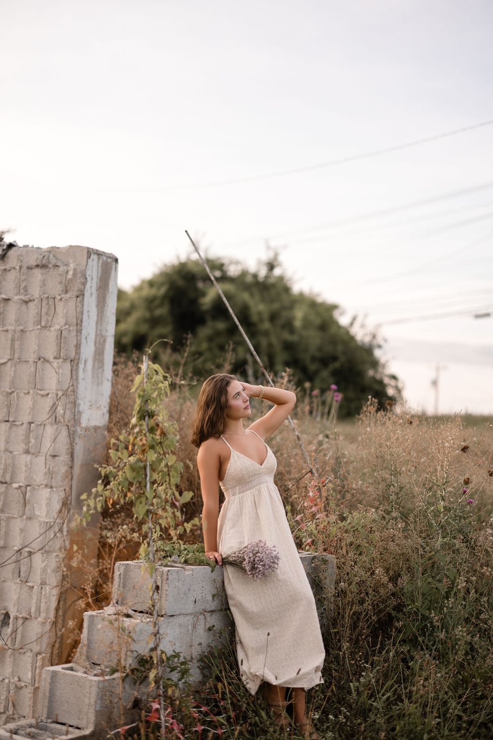 A person in a light-colored dress poses gracefully among concrete pillars and wild grass at sunset.