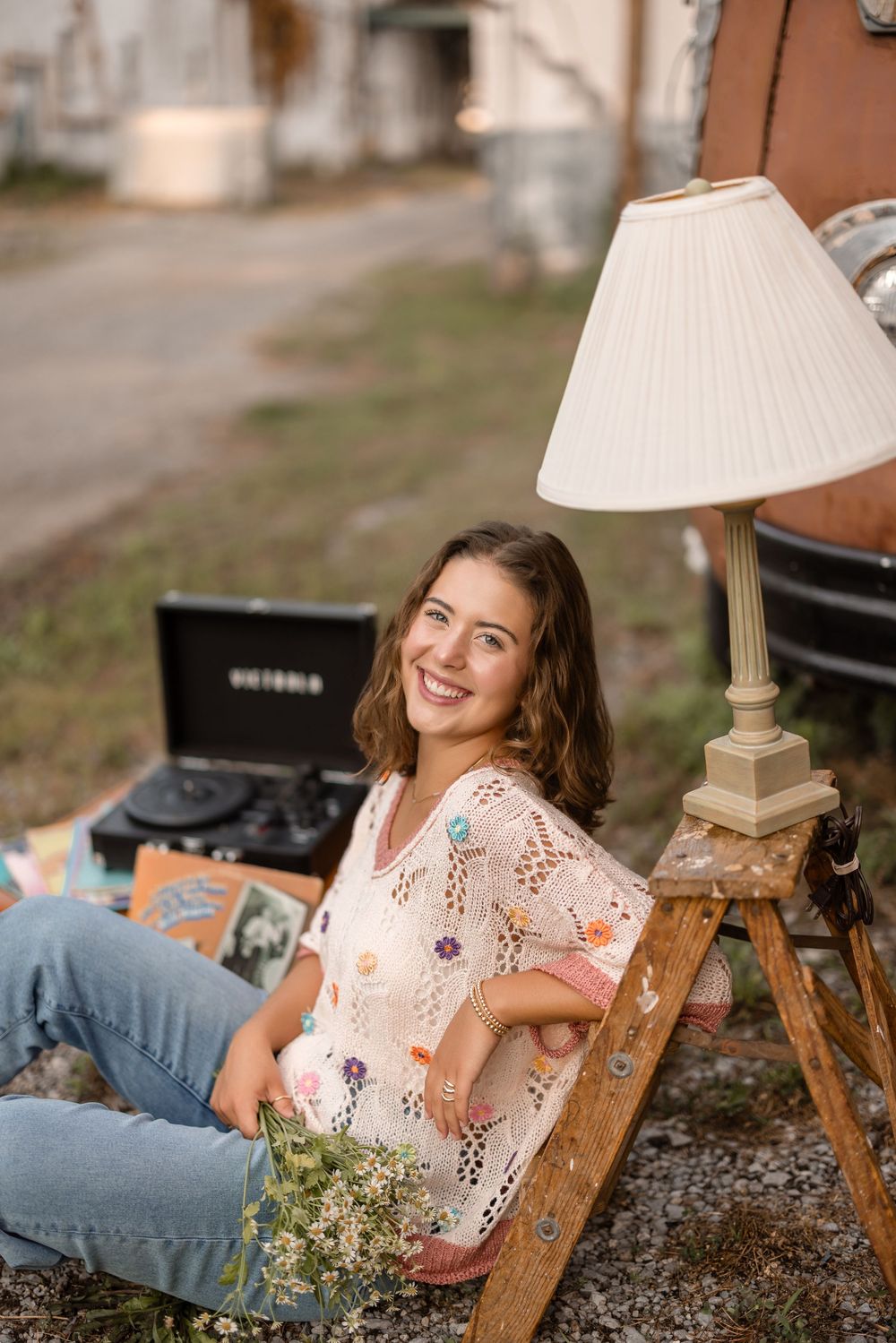 Woman in floral blouse sitting outdoors with vintage lamp and wooden ladder in rustic setting.