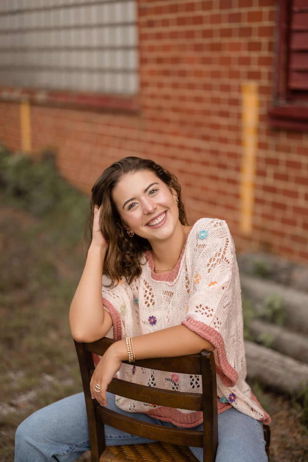 A person in a light lace top and jeans sits casually on a wooden chair against a brick wall backdrop.