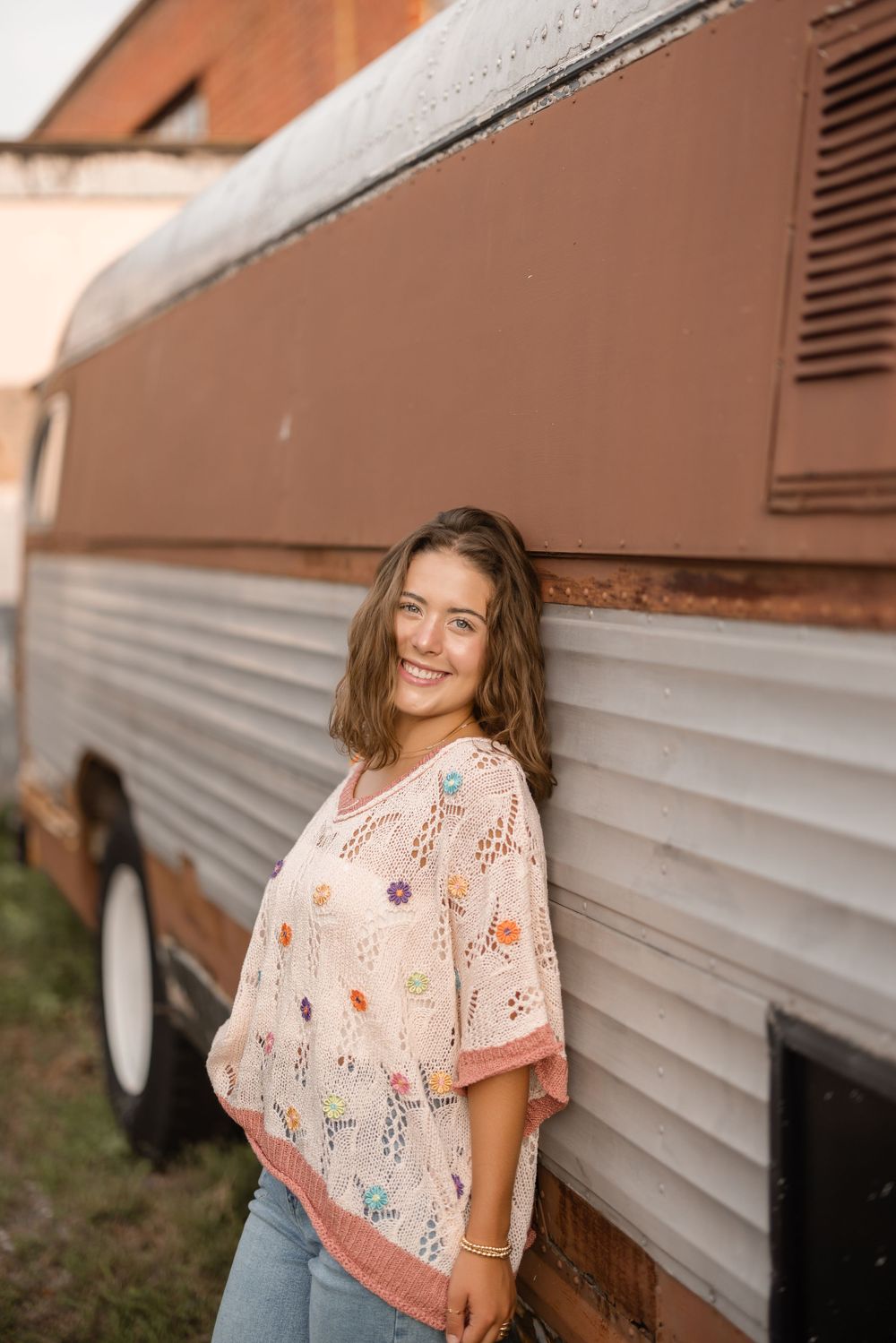 Someone wearing a floral print blouse and jeans stands smiling next to a vintage white and orange camper van.