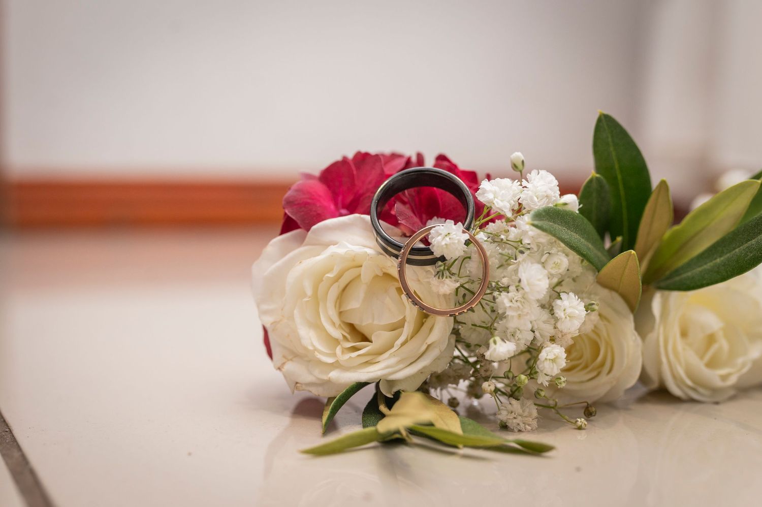 Wedding rings nestled among white roses, pink flowers and baby's breath on a light surface.