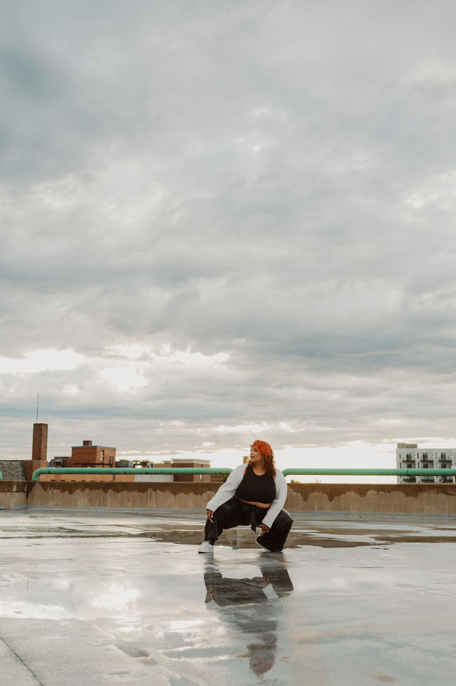 A curvy model poses on a rooftop wearing a black crop top and wide-leg pants against a cloudy sky.