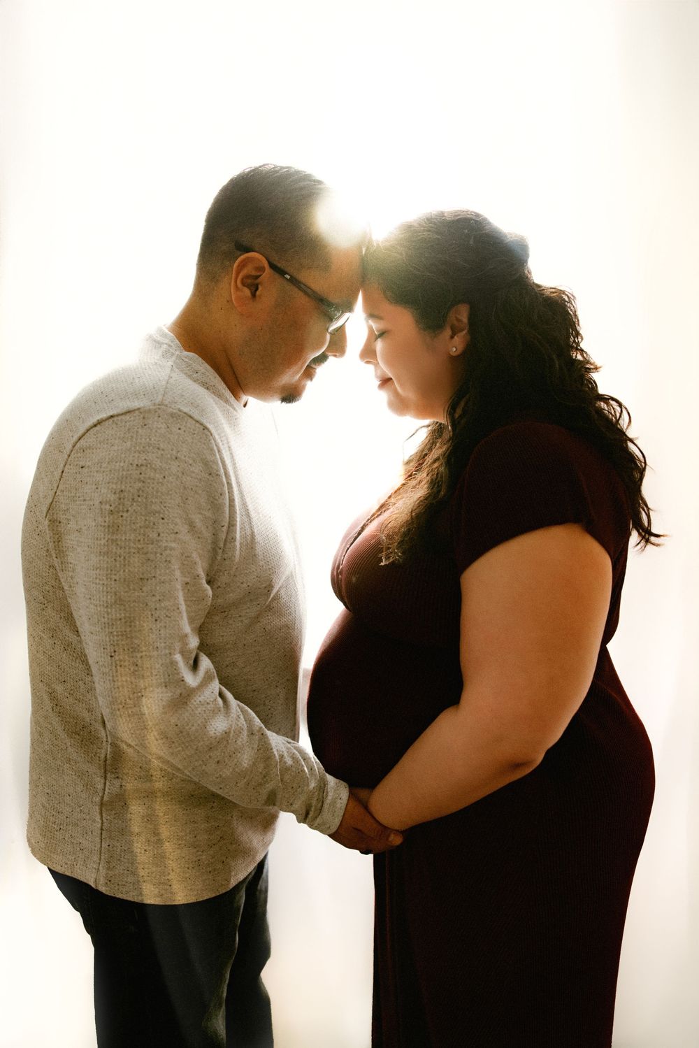 Silhouetted couple sharing intimate moment with pregnant belly against bright window.