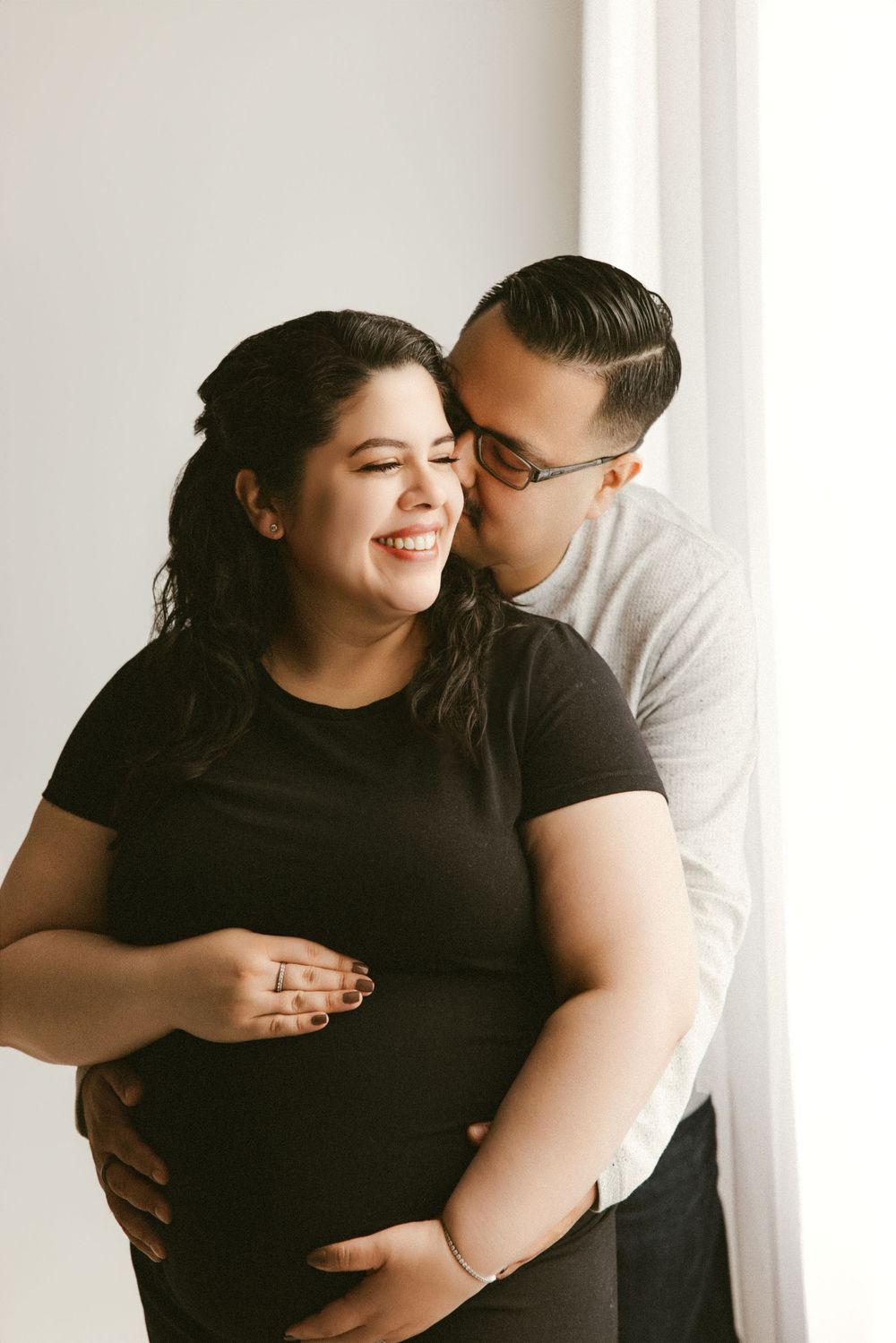 A loving maternity photo of an expecting couple embracing by a window in natural light.