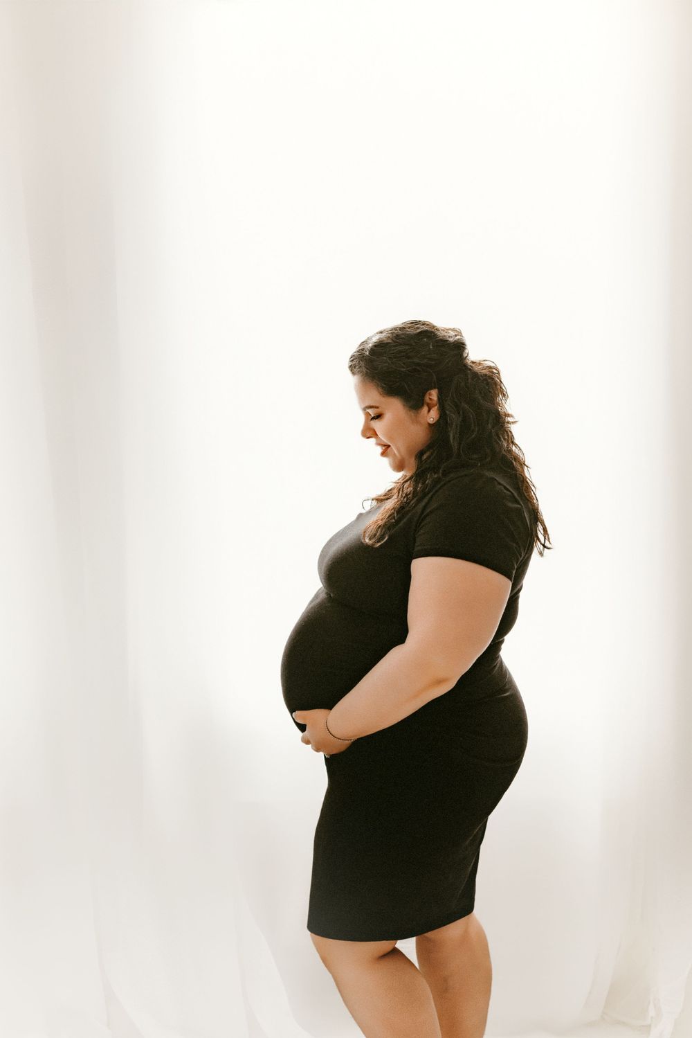 Side profile of pregnant woman in black dress smiling while cradling her baby bump.