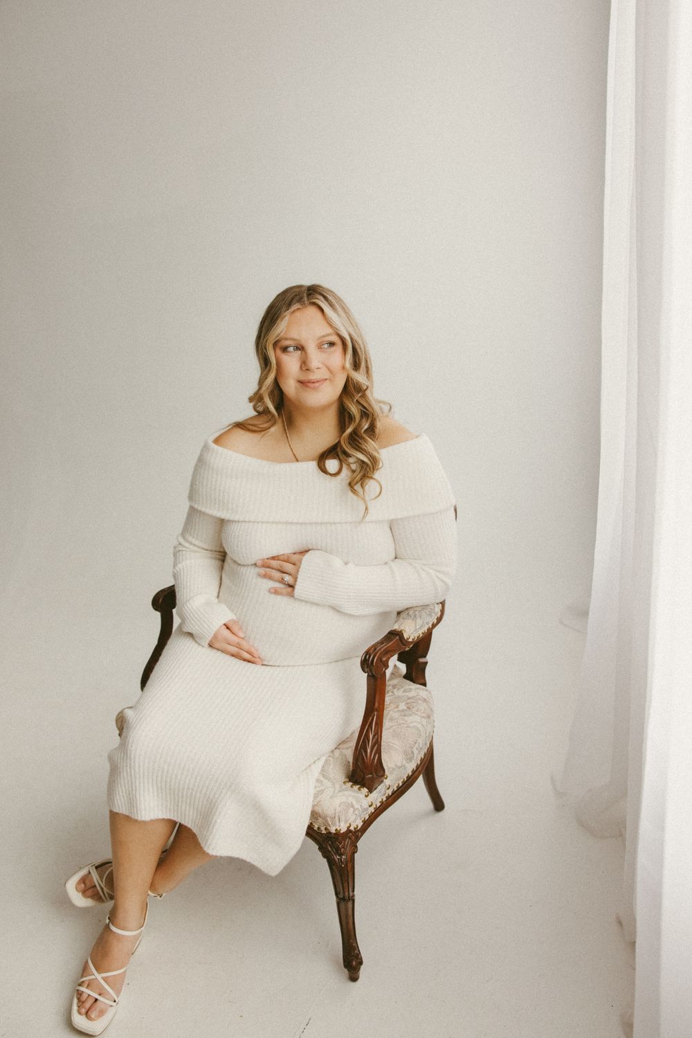 Expectant mother in white off-shoulder sweater dress poses elegantly on vintage chair.