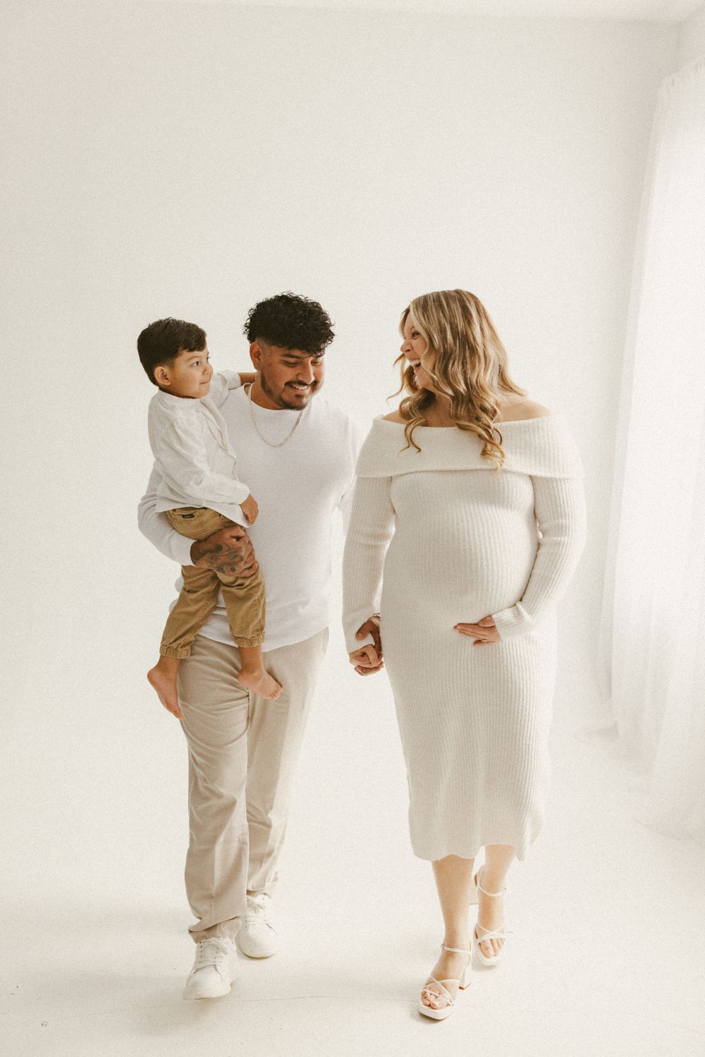 Family maternity photo with couple in white outfits and young child walking together.