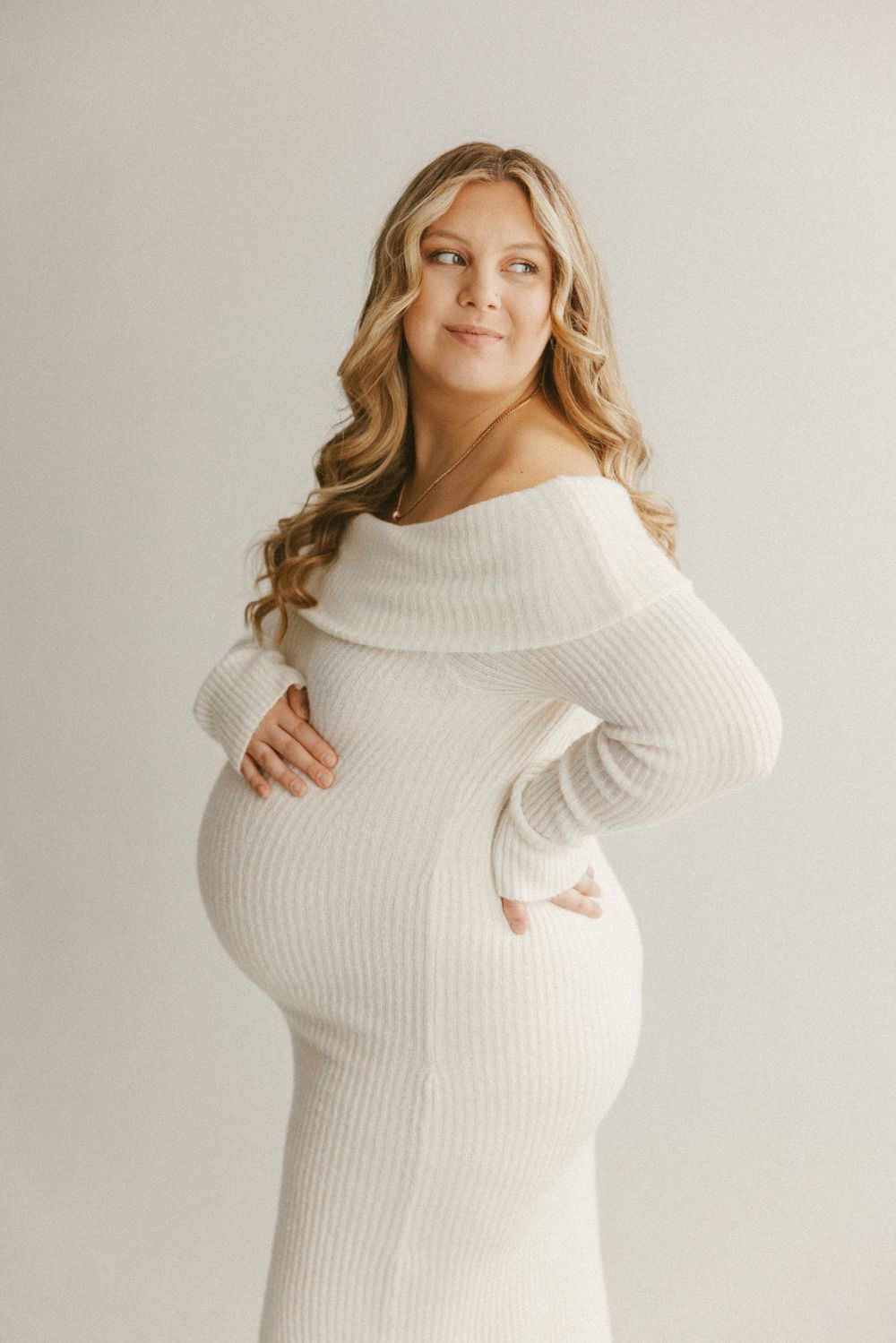 Pregnant woman in white ribbed sweater dress poses while holding her baby bump.