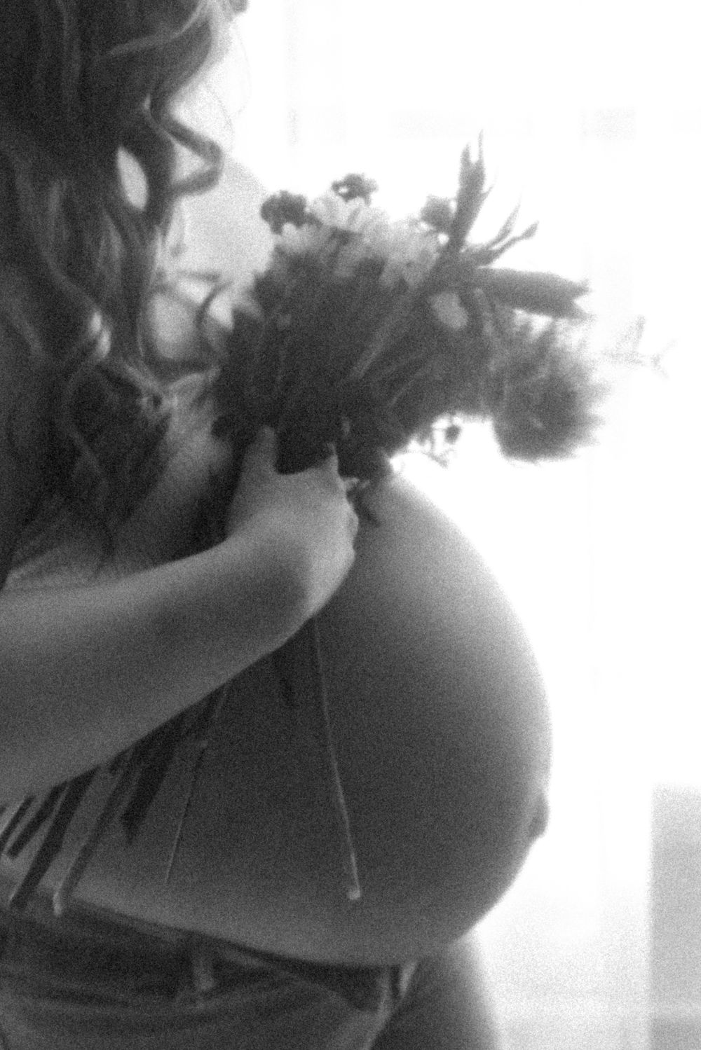 Artistic black and white photo of pregnant woman holding flowers against window light.