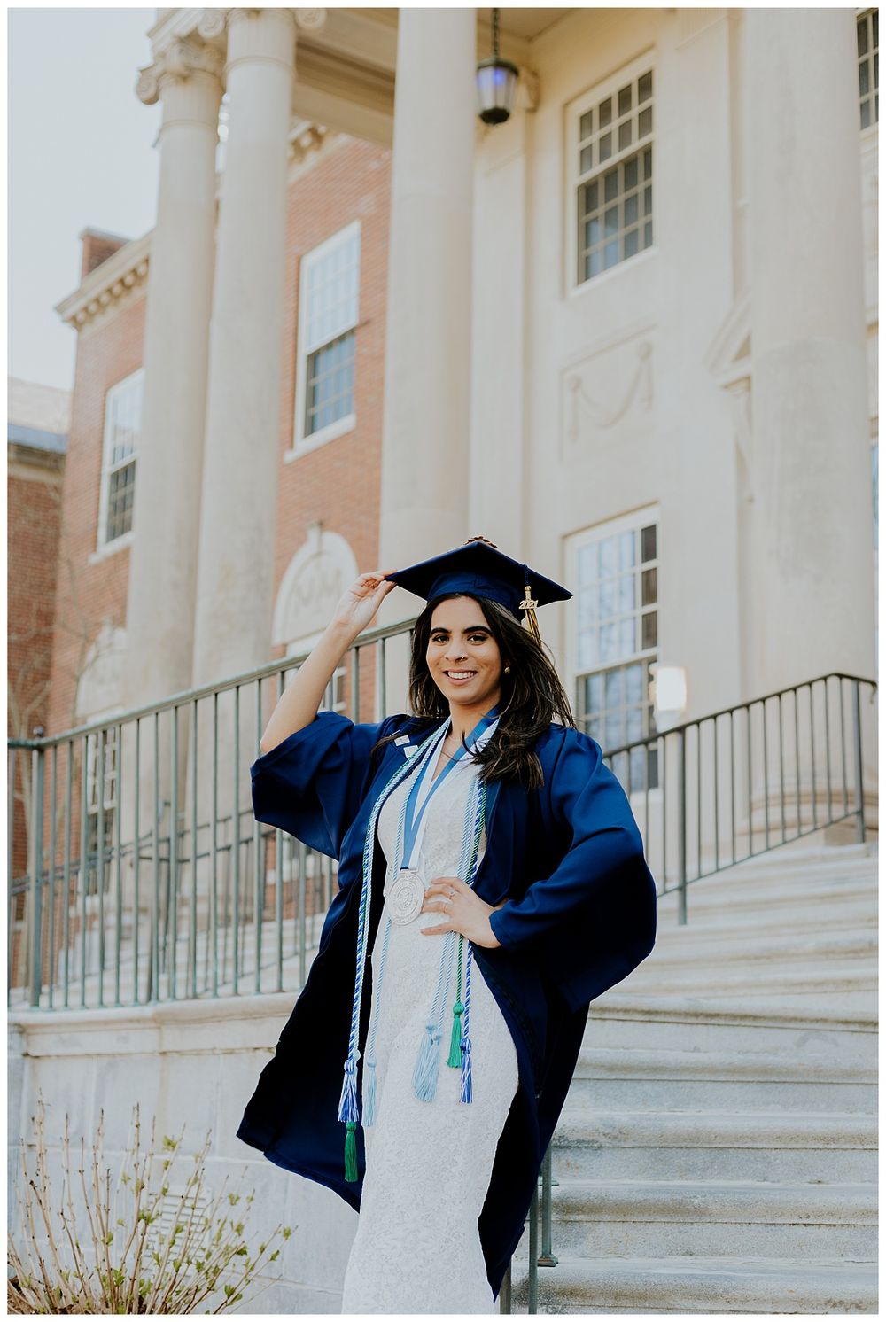 Graduate in blue cap and gown poses on white pillared building steps.