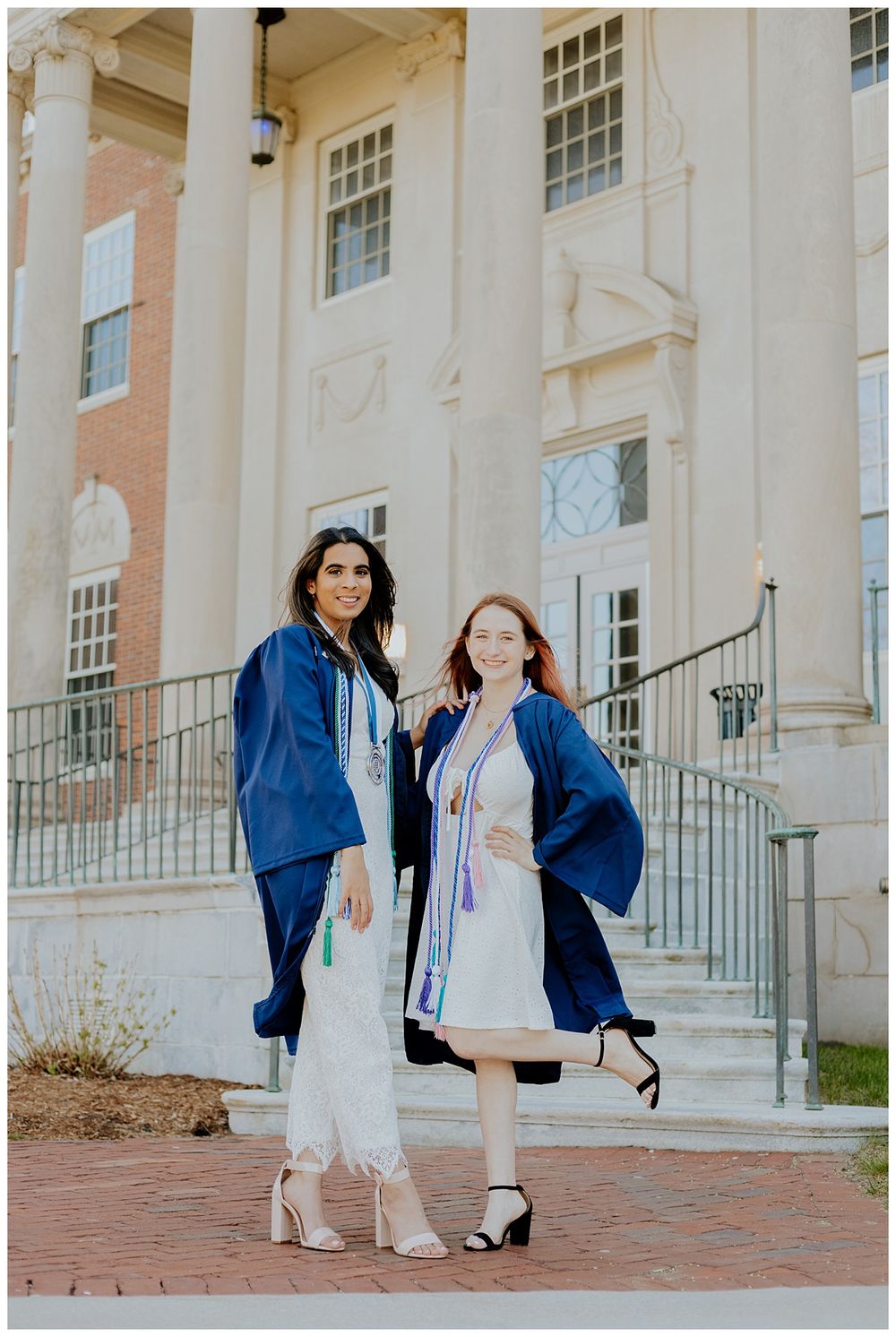 Two graduates in blue caps and gowns pose on steps of classical building.