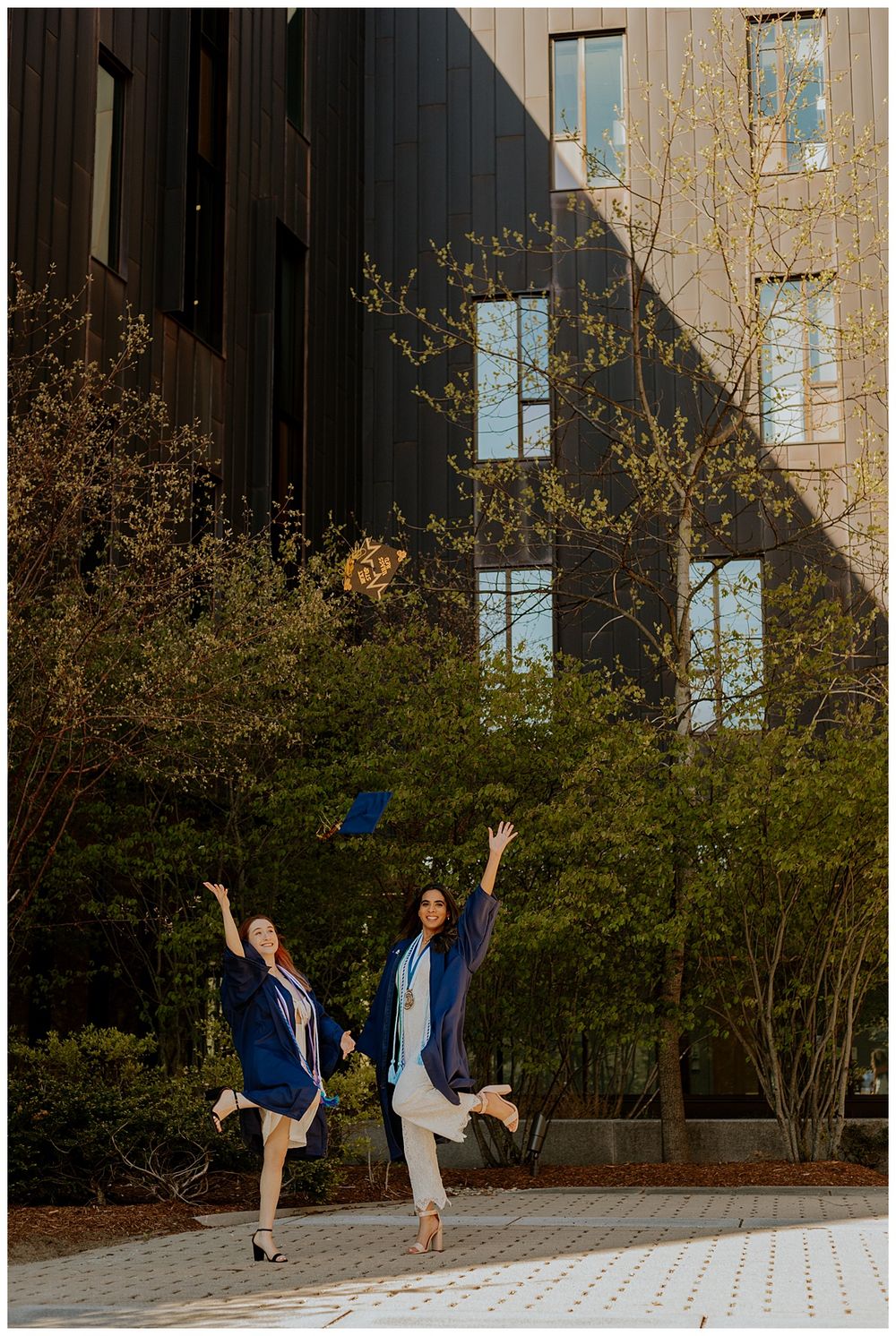 Two graduates in blue caps and gowns jump joyfully in front of a modern building with trees and bushes.
