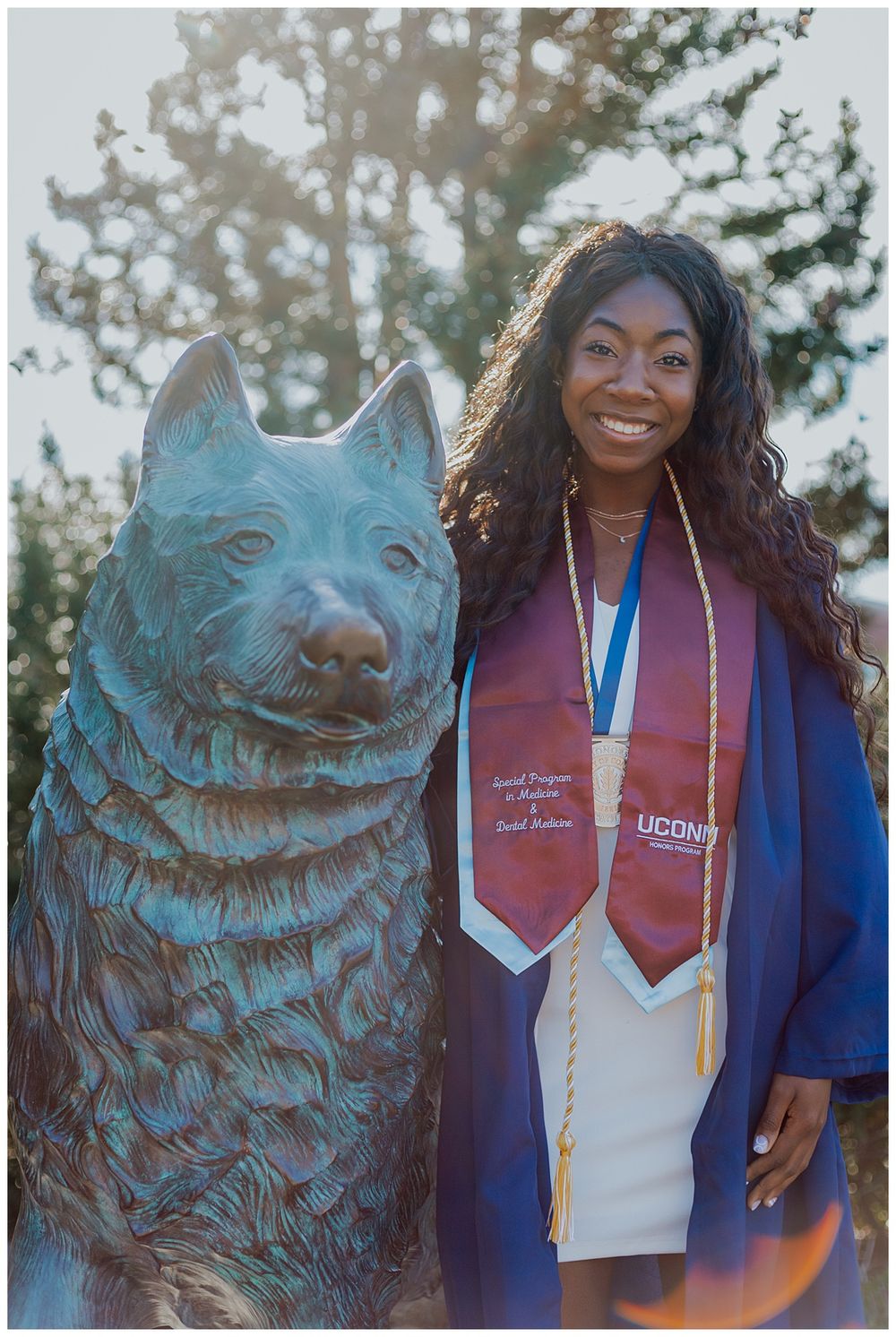 Graduate in commencement regalia poses with the iconic UConn Husky statue on campus.