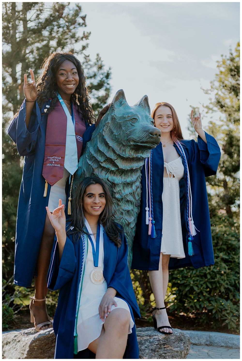 Three graduates in blue robes celebrate around the UConn Husky statue during graduation.