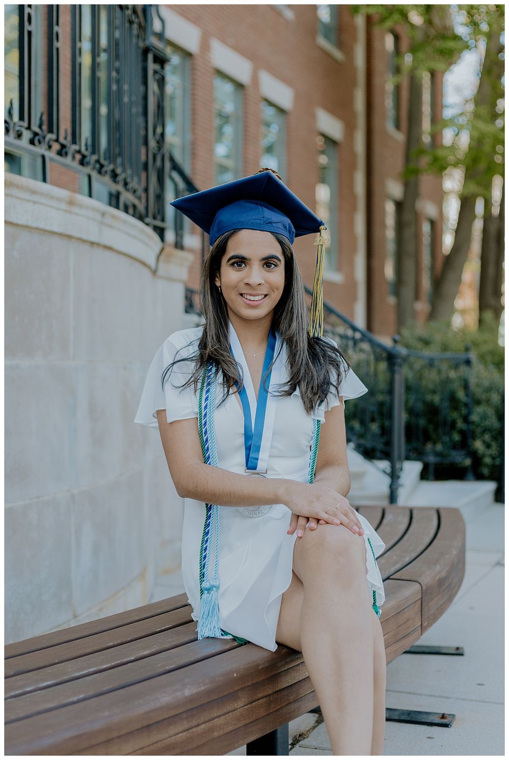 Graduate in blue cap and white dress poses on wooden bench during commencement.