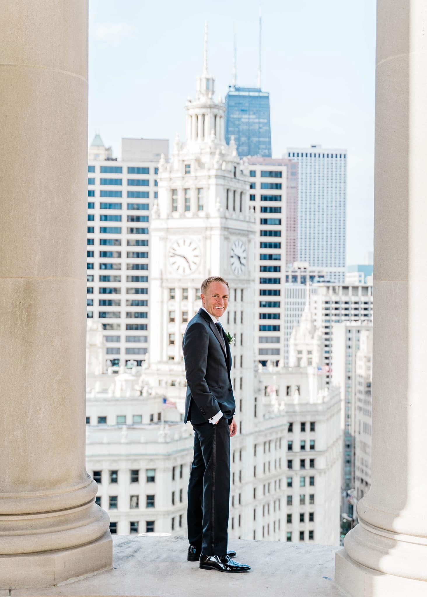 Man in black suit stands between columns overlooking historic Chicago Wrigley Building clock tower and downtown skyline.