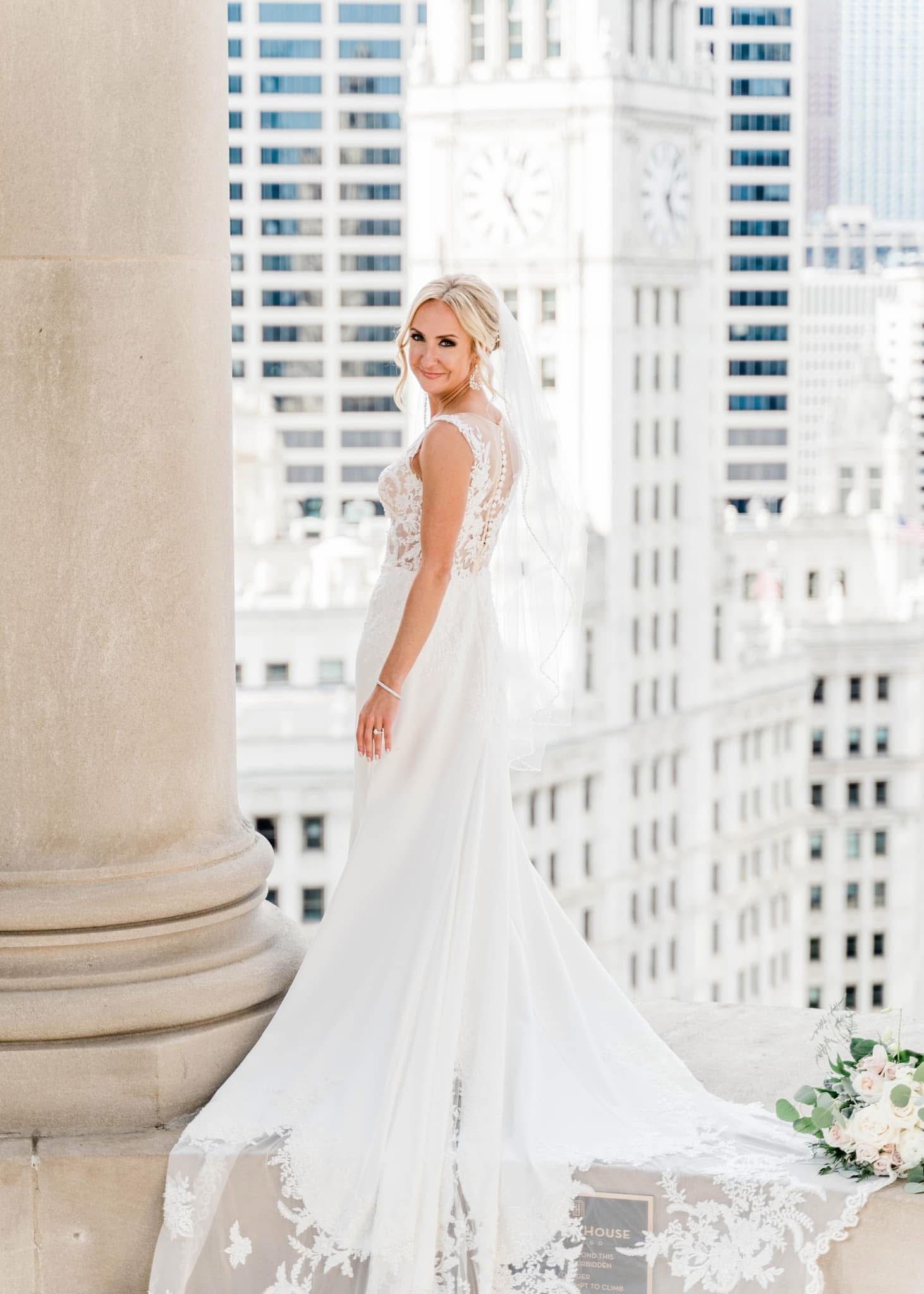 Elegant wedding dress with lace details and flowing train photographed against Chicago cityscape from historic building balcony.
