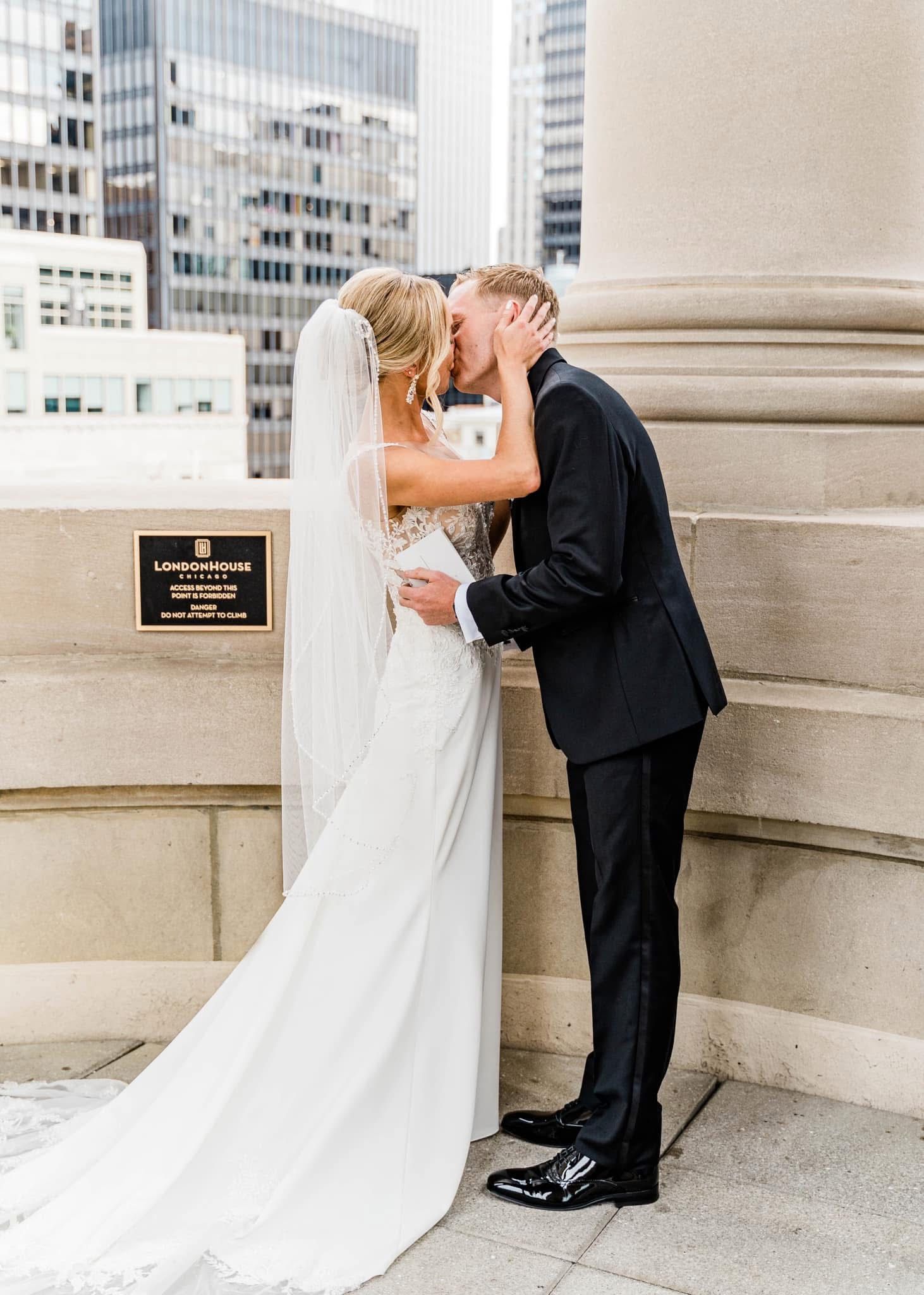 A bride and groom share their first kiss as newlyweds on a rooftop terrace with city buildings in the background.