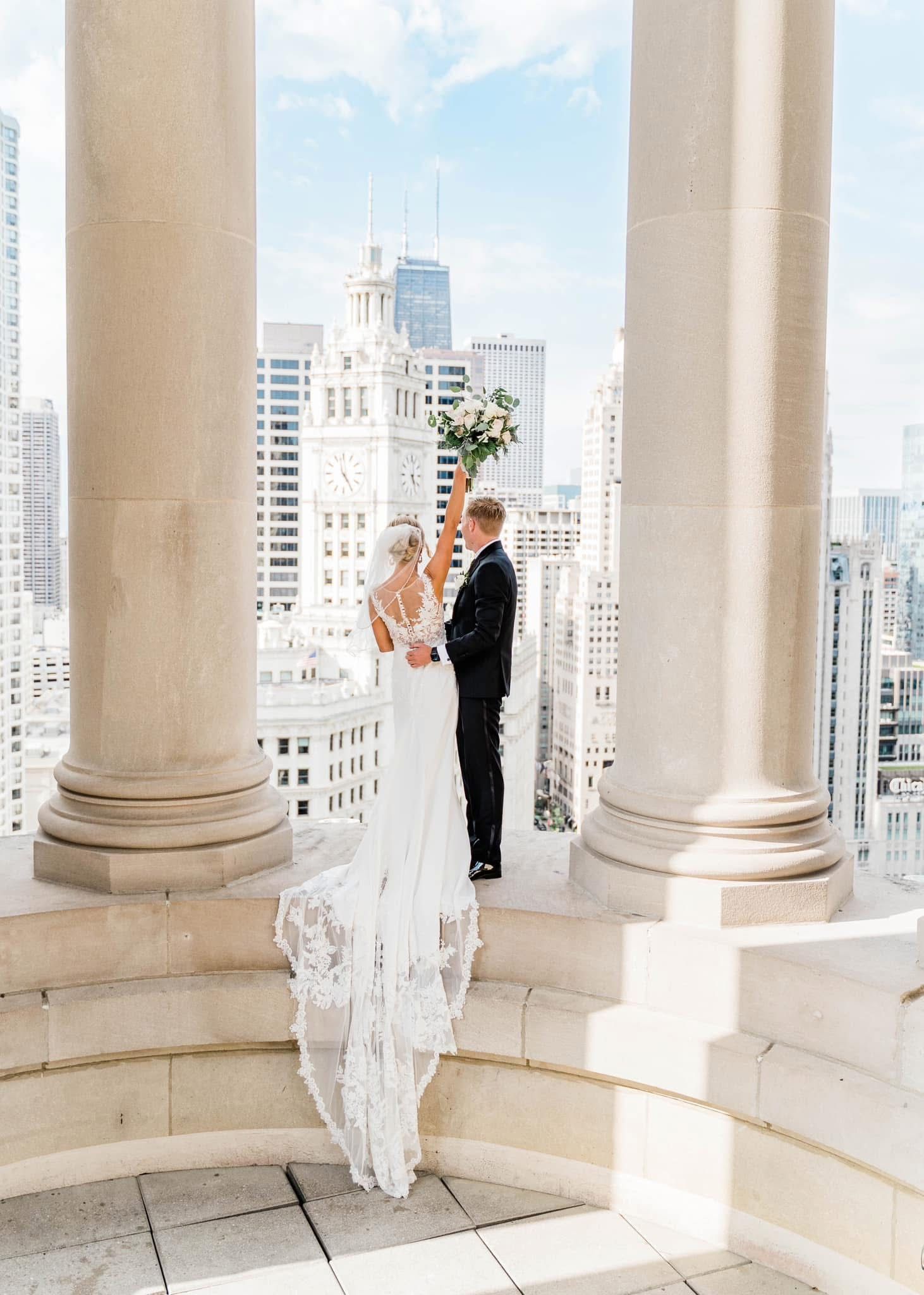 A wedding couple shares a romantic moment between elegant white columns overlooking a city skyline.
