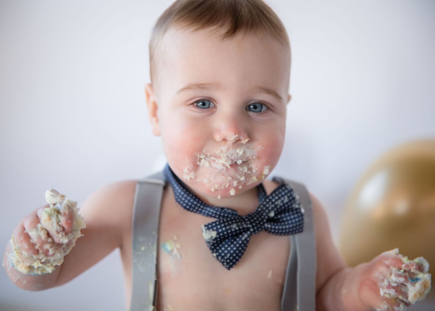 A small child wearing suspenders and a bowtie enjoys smashing cake with frosting covering their face during a celebration.