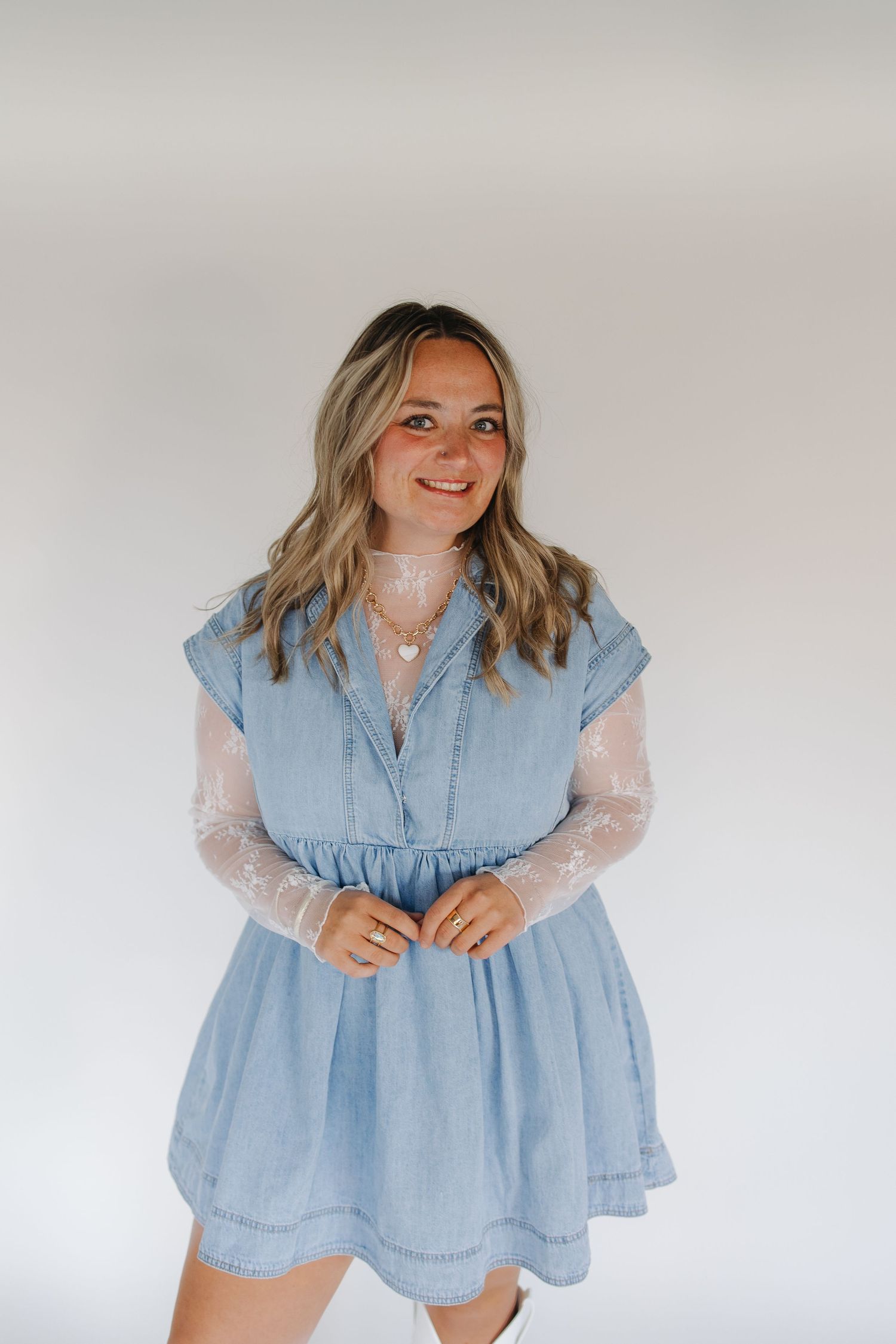 Person wearing light blue denim dress with sheer white long sleeves poses against neutral background.
