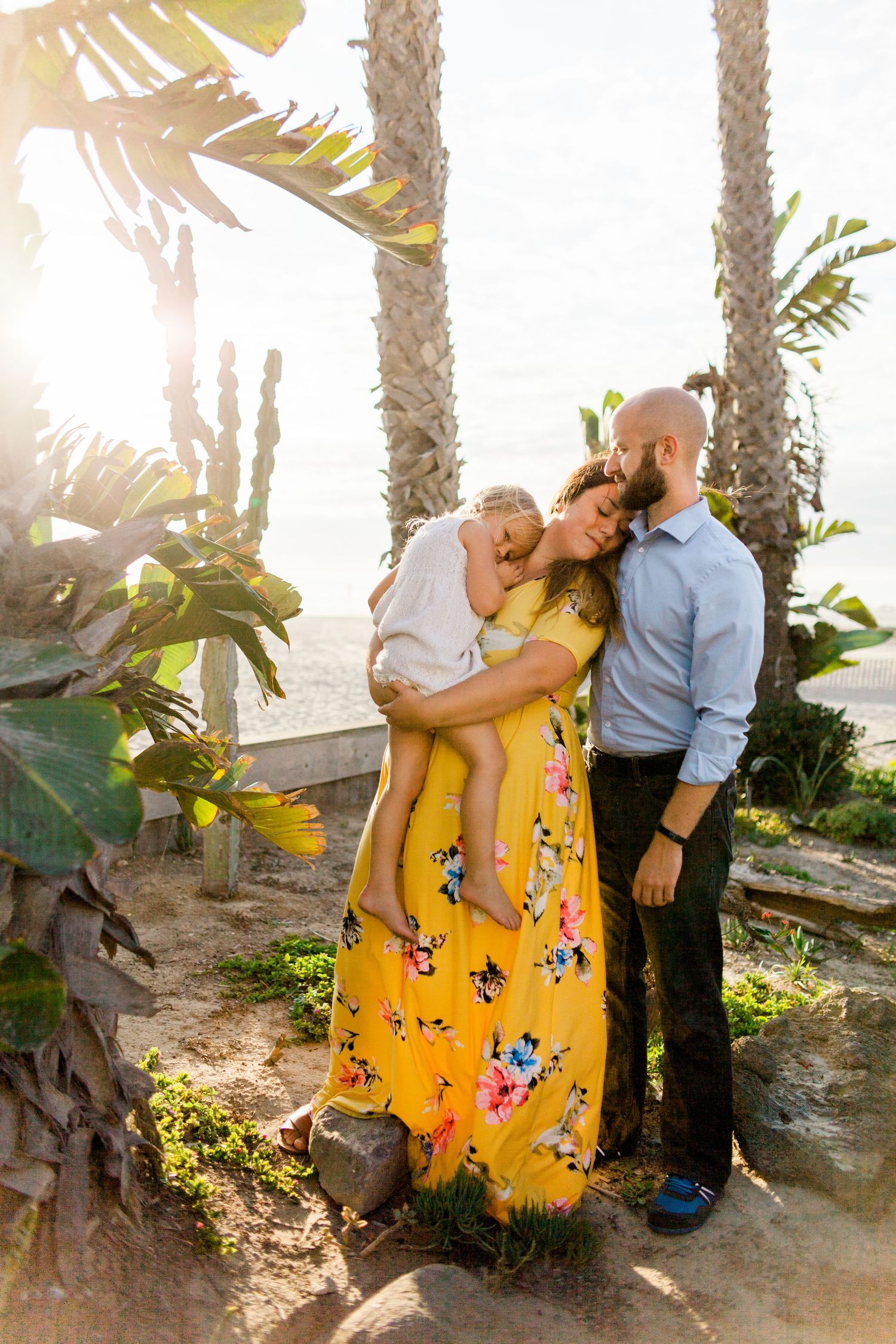 mom in a yellow dress holding little girl in a white dress and leaning her head against dad in a blue shirt