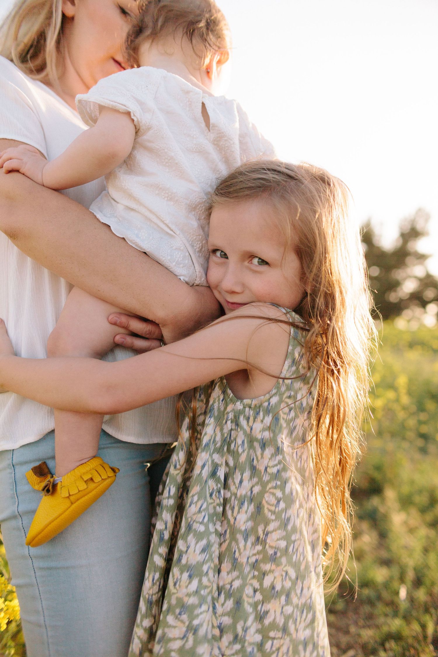 little girl hugging her mom and baby sister while looking at the camera