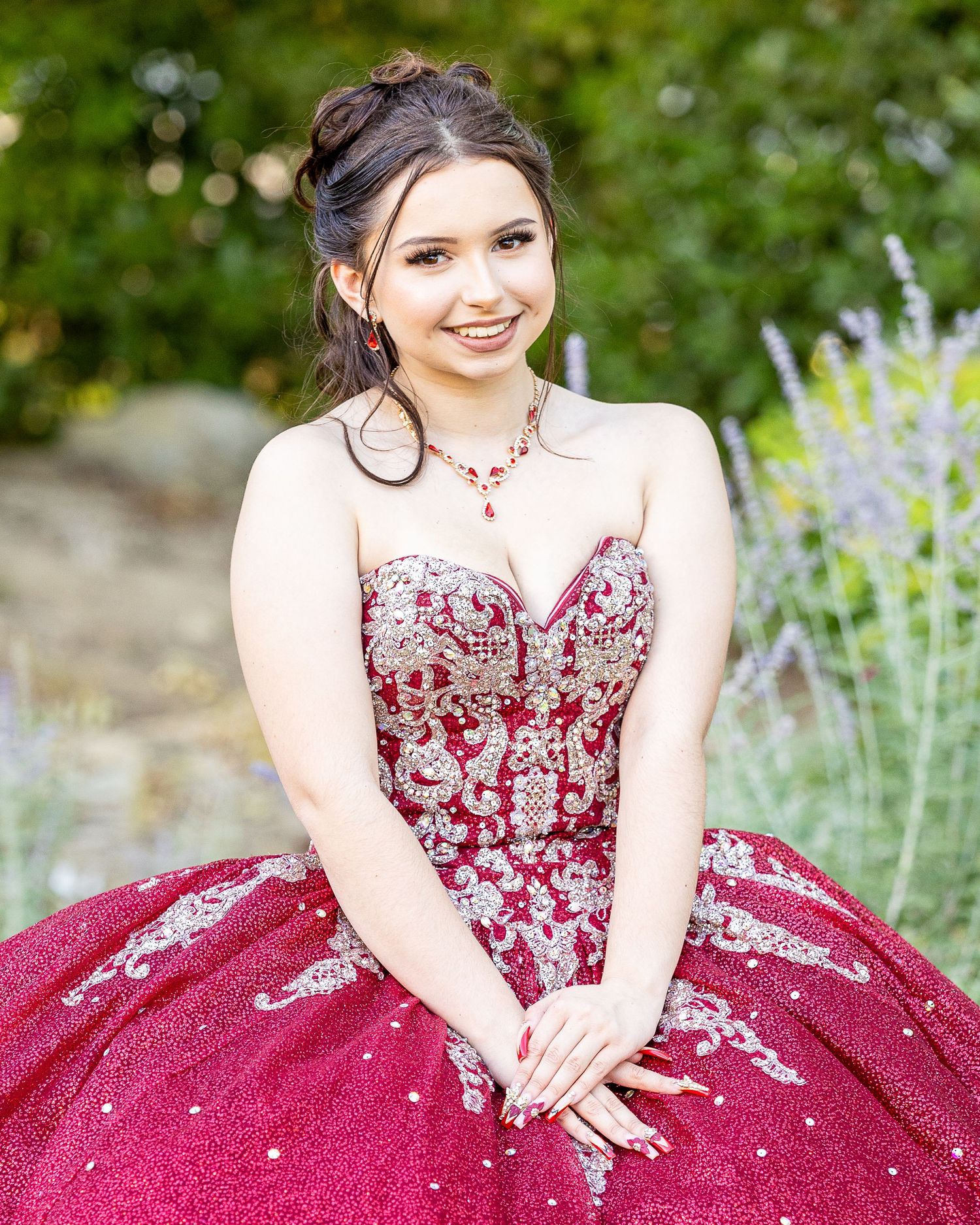 A person in a sparkling burgundy formal gown poses gracefully in a garden setting with lavender flowers.