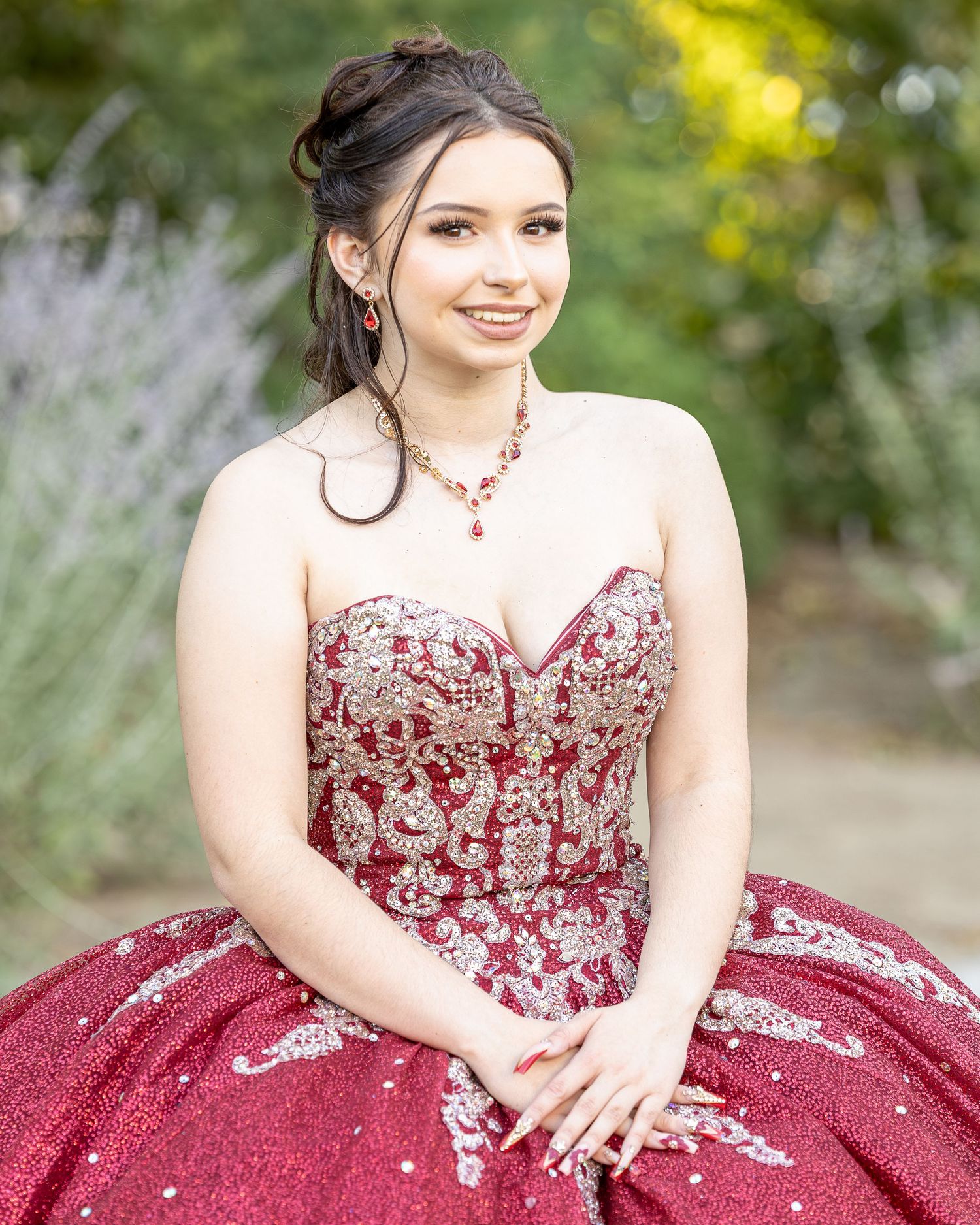 A woman in a strapless burgundy ball gown with silver embellishments poses outdoors amid soft-focus greenery.