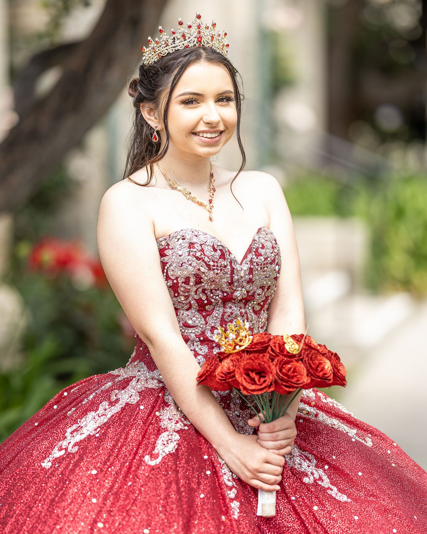 Beautiful red quinceanera dress with white lace details and crystal tiara, holding a bouquet of red roses in garden setting.