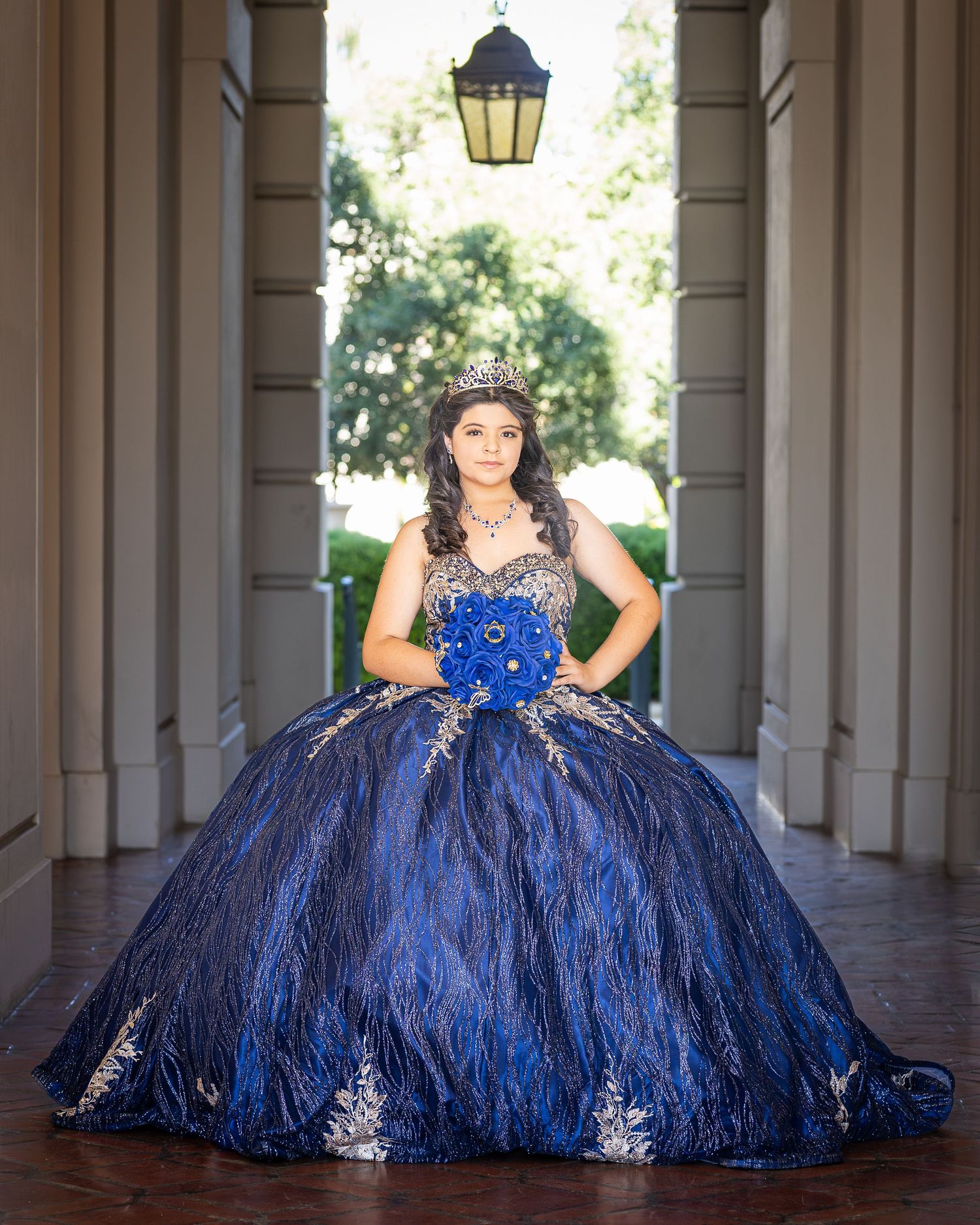 A royal blue quinceañera ball gown with silver embellishments is photographed under an elegant covered walkway.