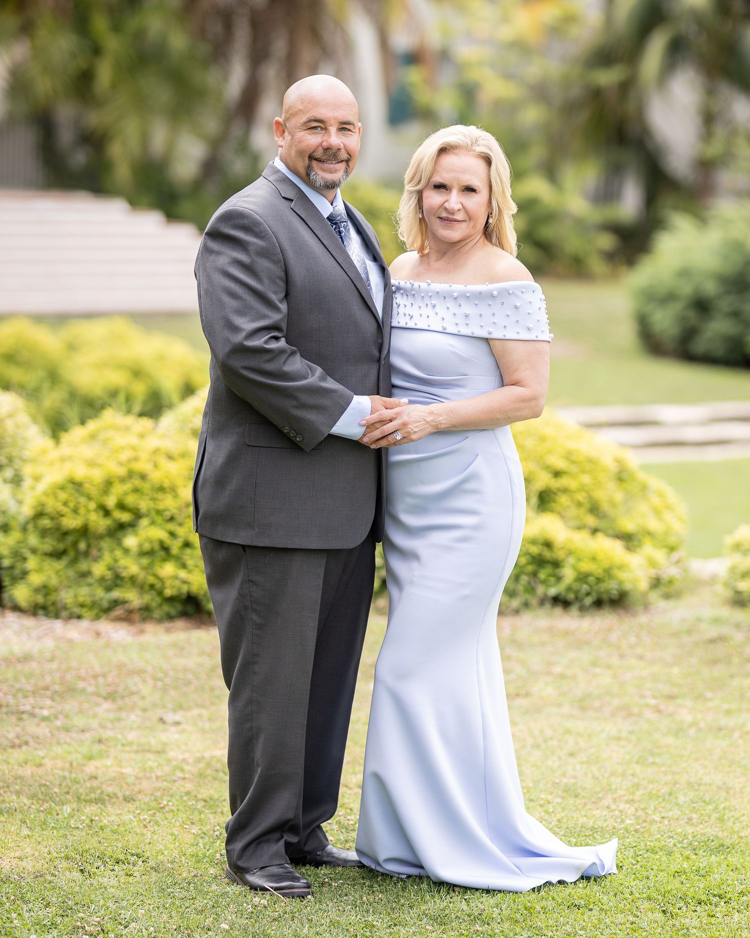 Wedding couple poses together on a grassy lawn with green shrubbery in the background.