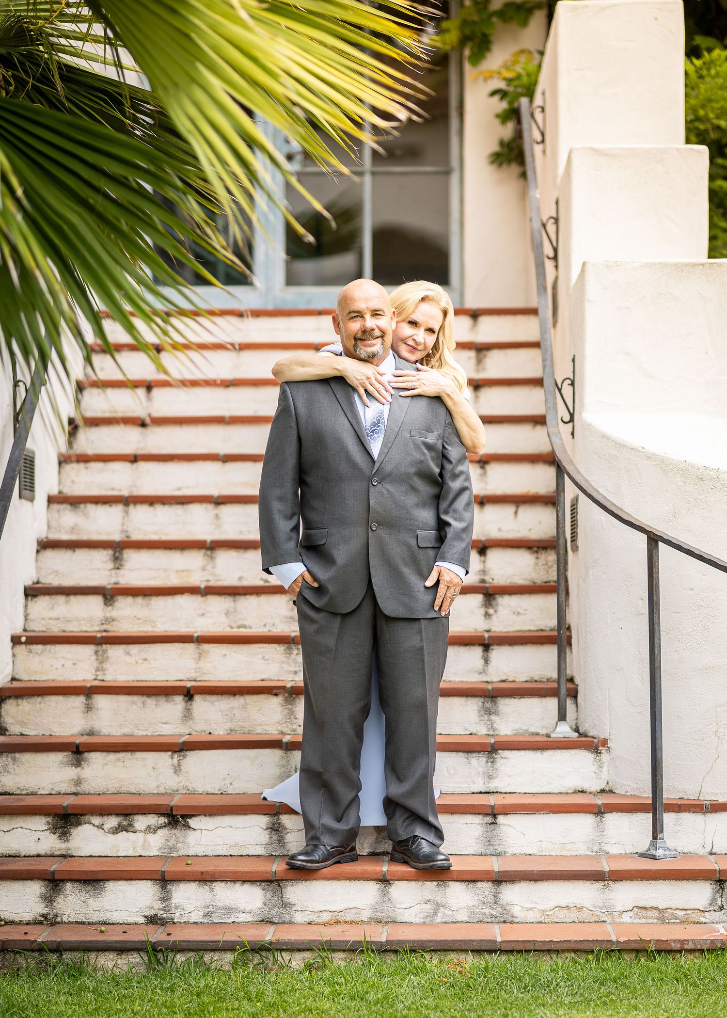 A wedding couple in formal attire stands together on brick steps with palm fronds visible in the background.
