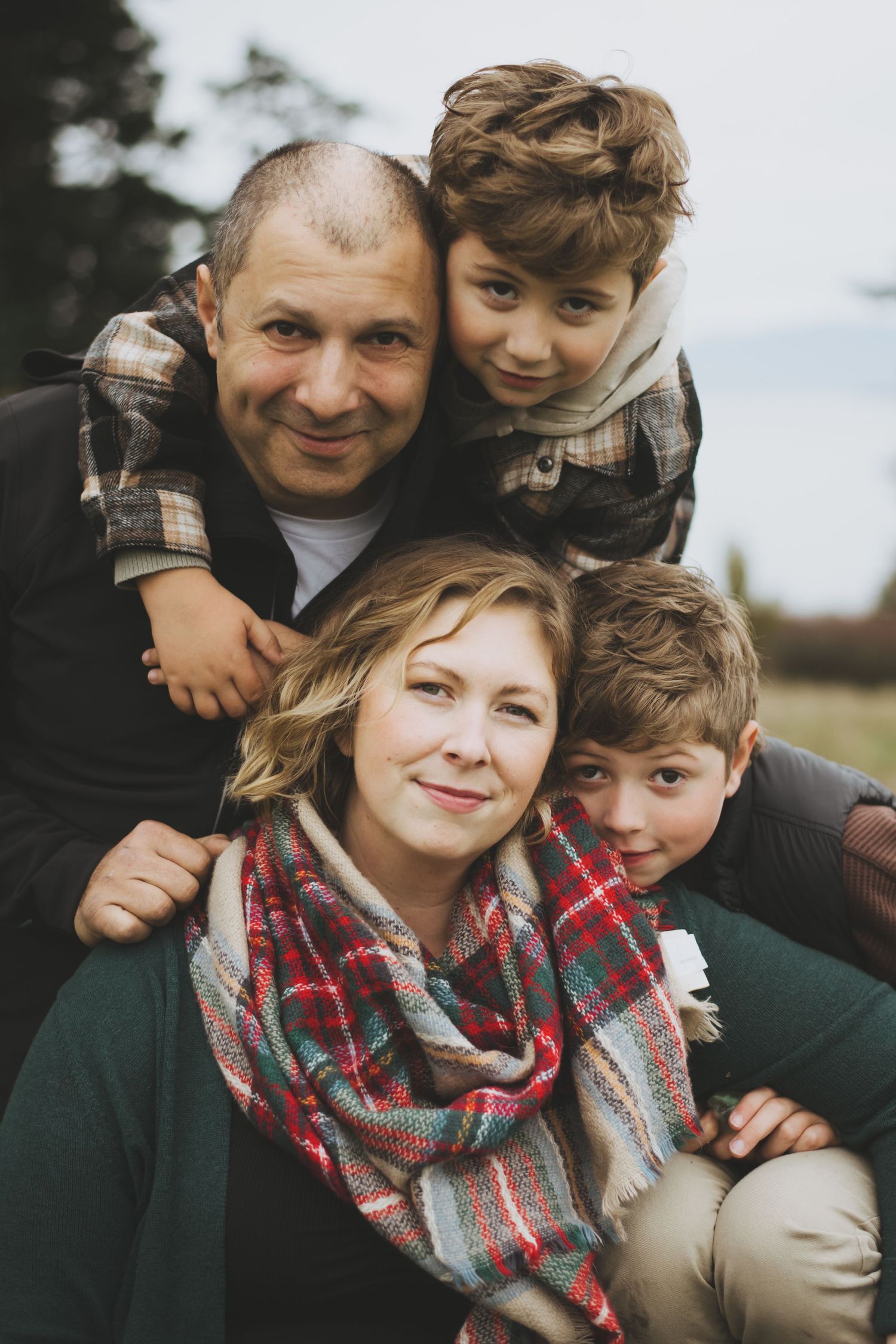 A family poses together outdoors wearing cozy fall clothing including a plaid shirt and patterned scarf.