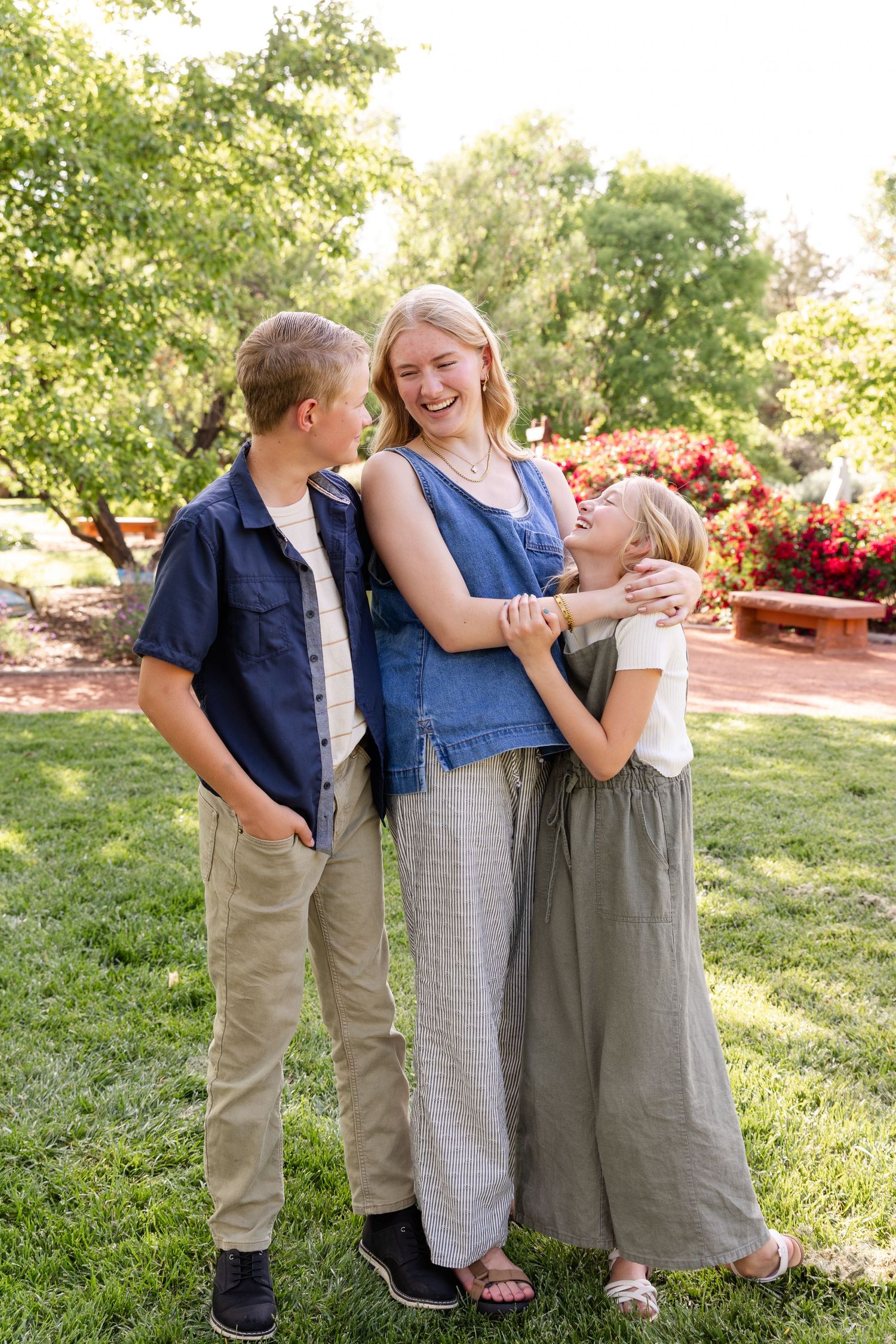 a teenage daughter in the middle looking to her right at a 14 year old brother laughing while hugging a 9 year old sister who is looking up