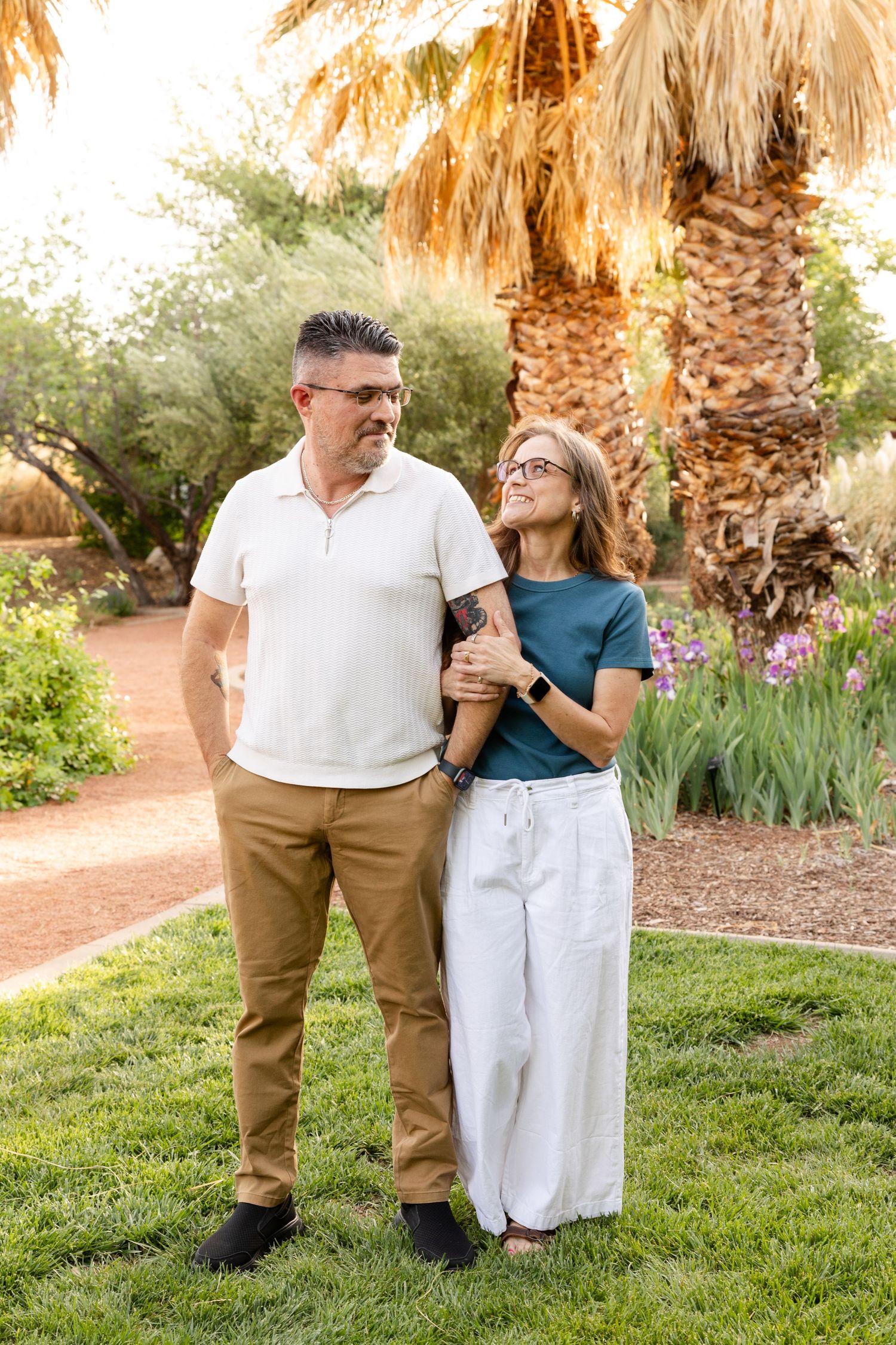 a husband and wife smiling at each other. standing in a grass with palm trees in the background