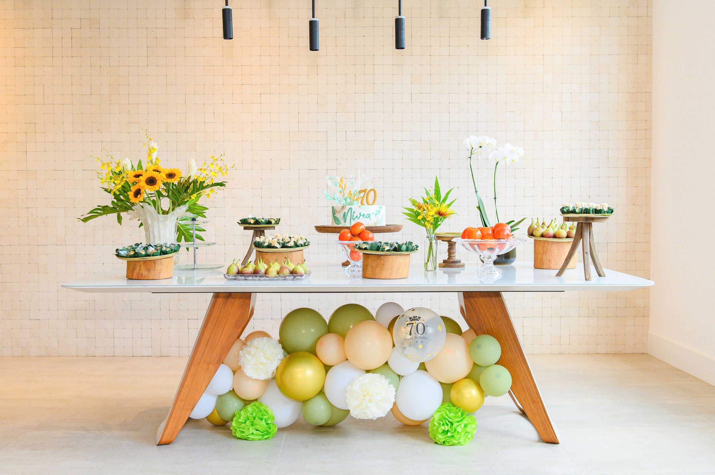 A modern dessert table with treats on elevated stands, decorated with green and white balloon garland underneath.
