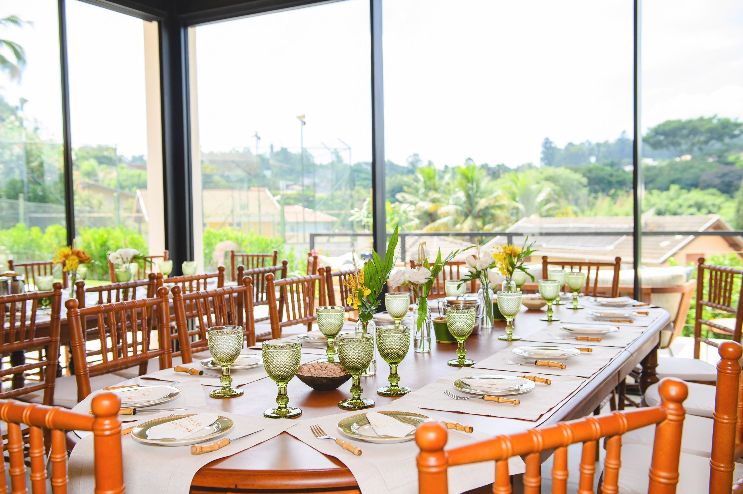 Long dining table set with glassware and centerpieces in an elegant venue with floor-to-ceiling windows overlooking greenery.
