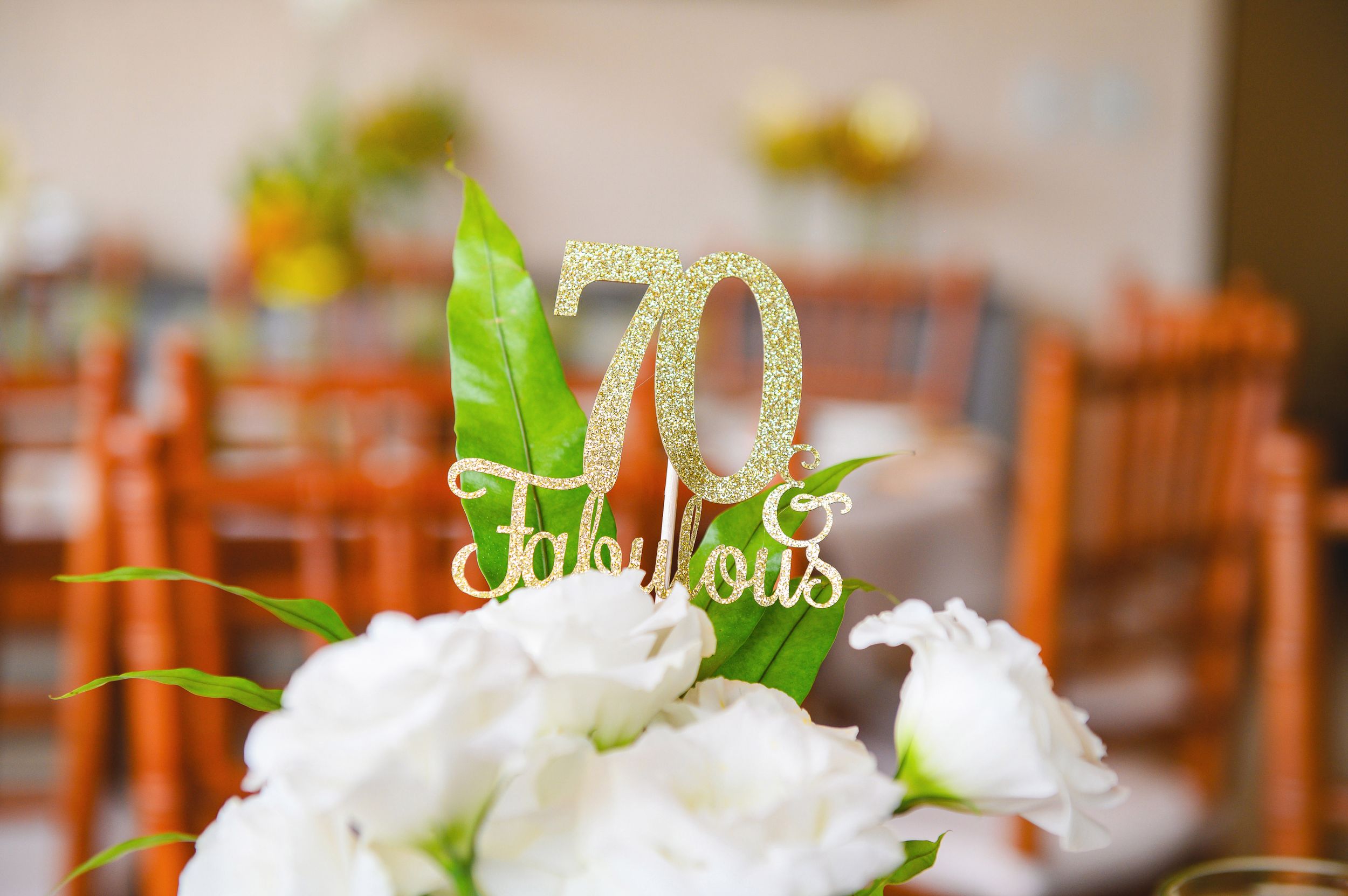 Gold number 70 table topper decoration on white floral centerpiece with blurred wooden chairs in background.