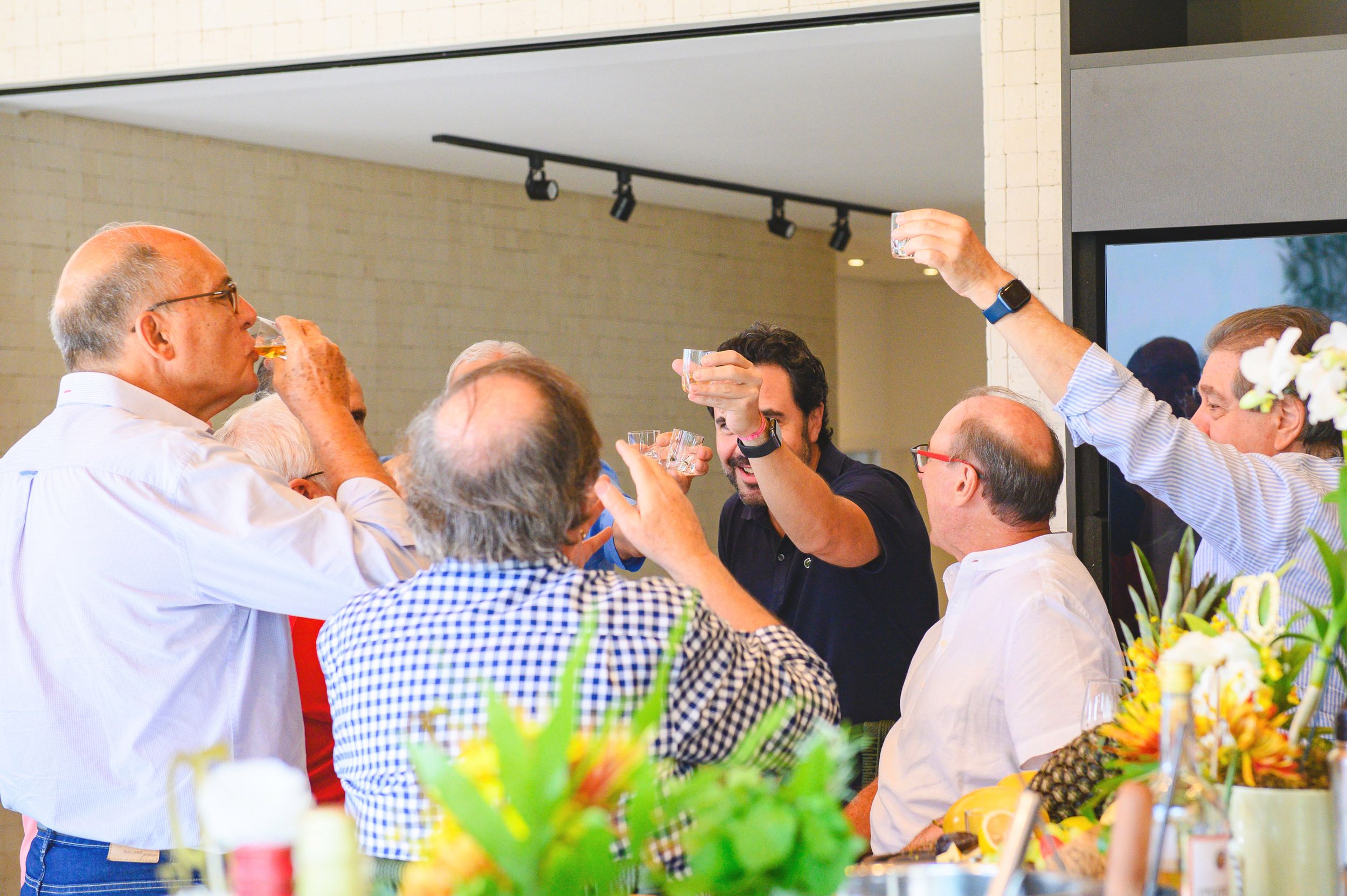A group of people raise their glasses in a toast at an indoor gathering with flowers and decor visible.