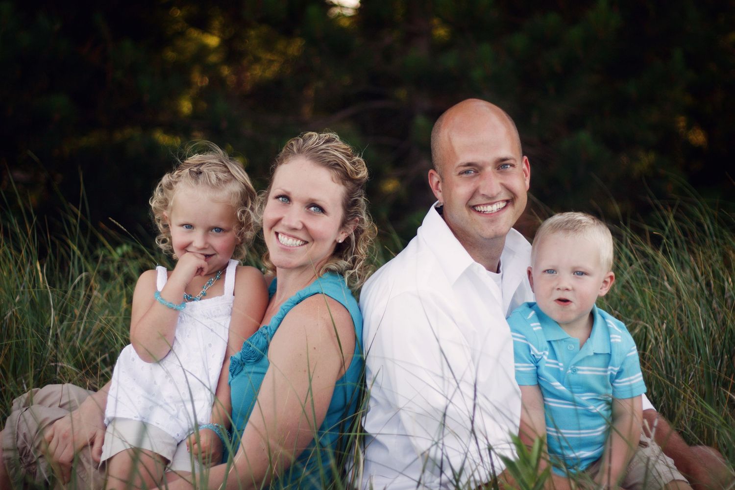 A family of four sits together outdoors in a natural setting with tall grass and trees in the background.